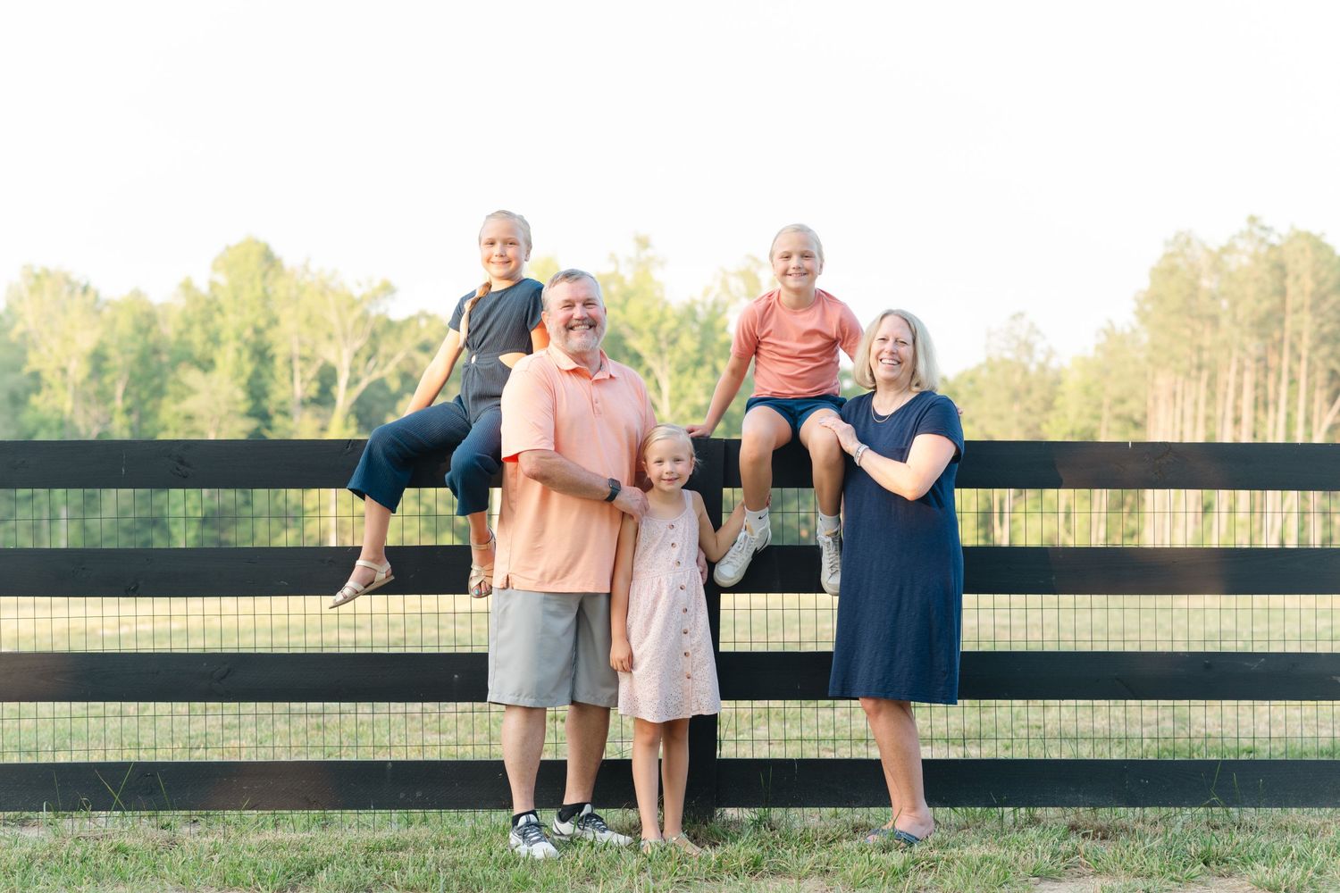 Family of five posing by a black wooden fence outdoors, with two kids sitting on top and one standing between parents.