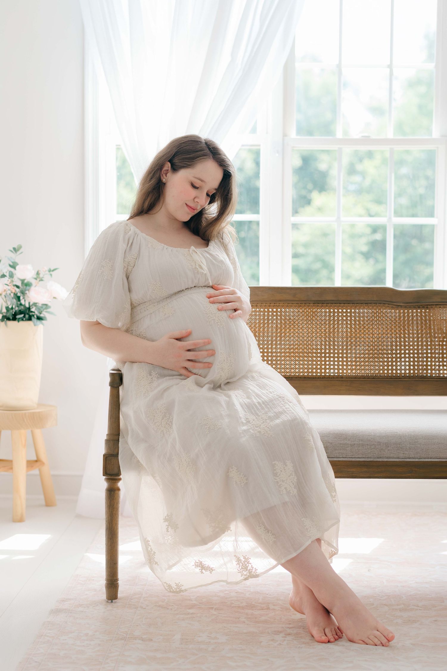 Pregnant woman in white lace dress sitting barefoot on chair, cradling baby bump near sunlit window.
