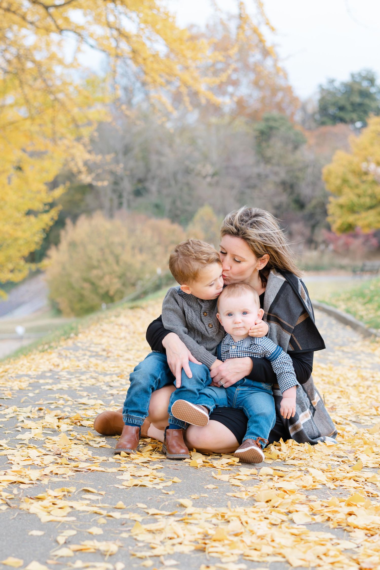 Mother kissing older son while hugging both boys on leaf-covered path in autumn park.