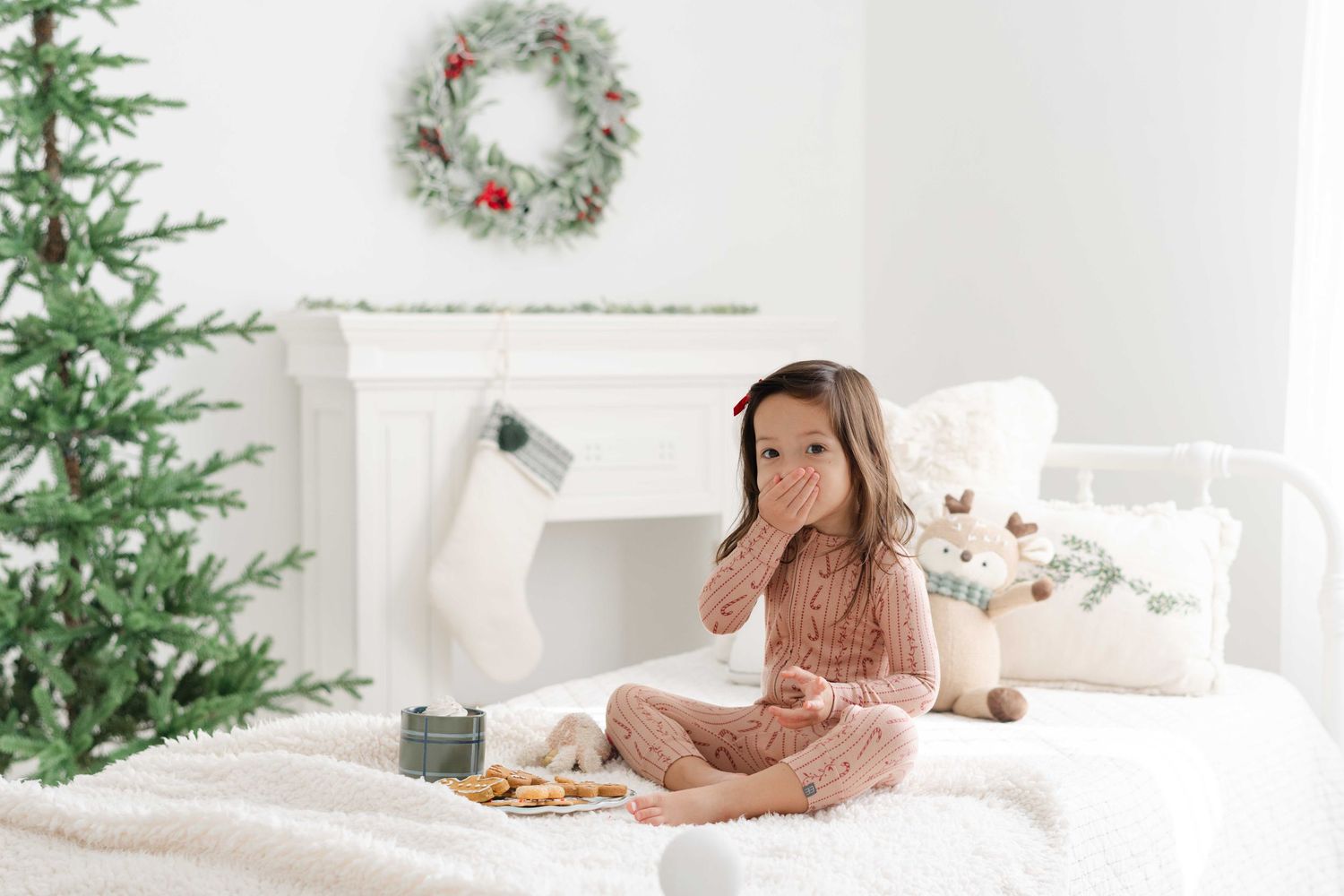 Young girl in pink pajamas sitting on white bed with Christmas cookies, tree, and fireplace with stocking.