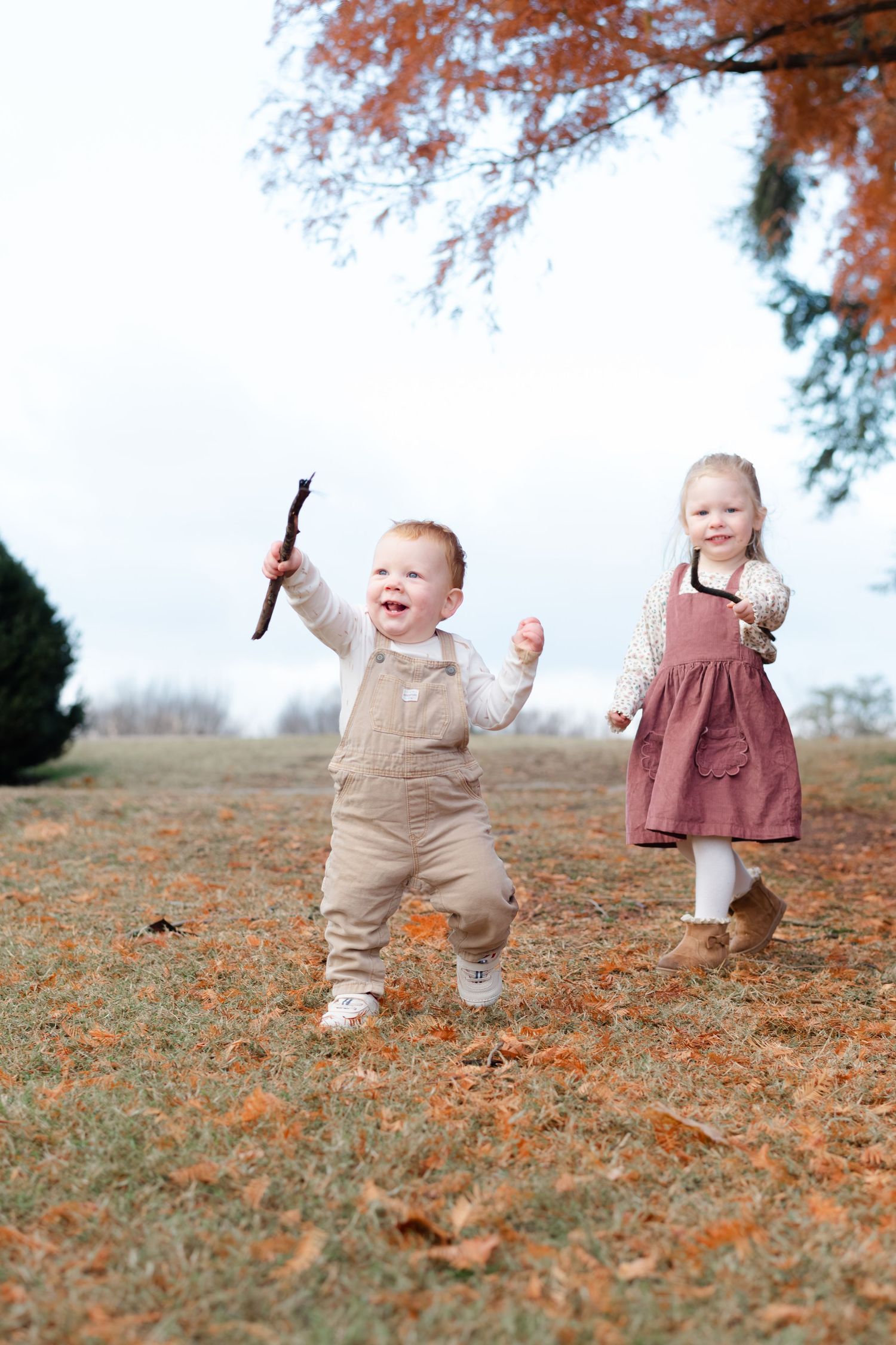 Two toddlers playing outdoors in autumn, one holding a stick in overalls, the other in a pink dress, running on leaf-covered grass.