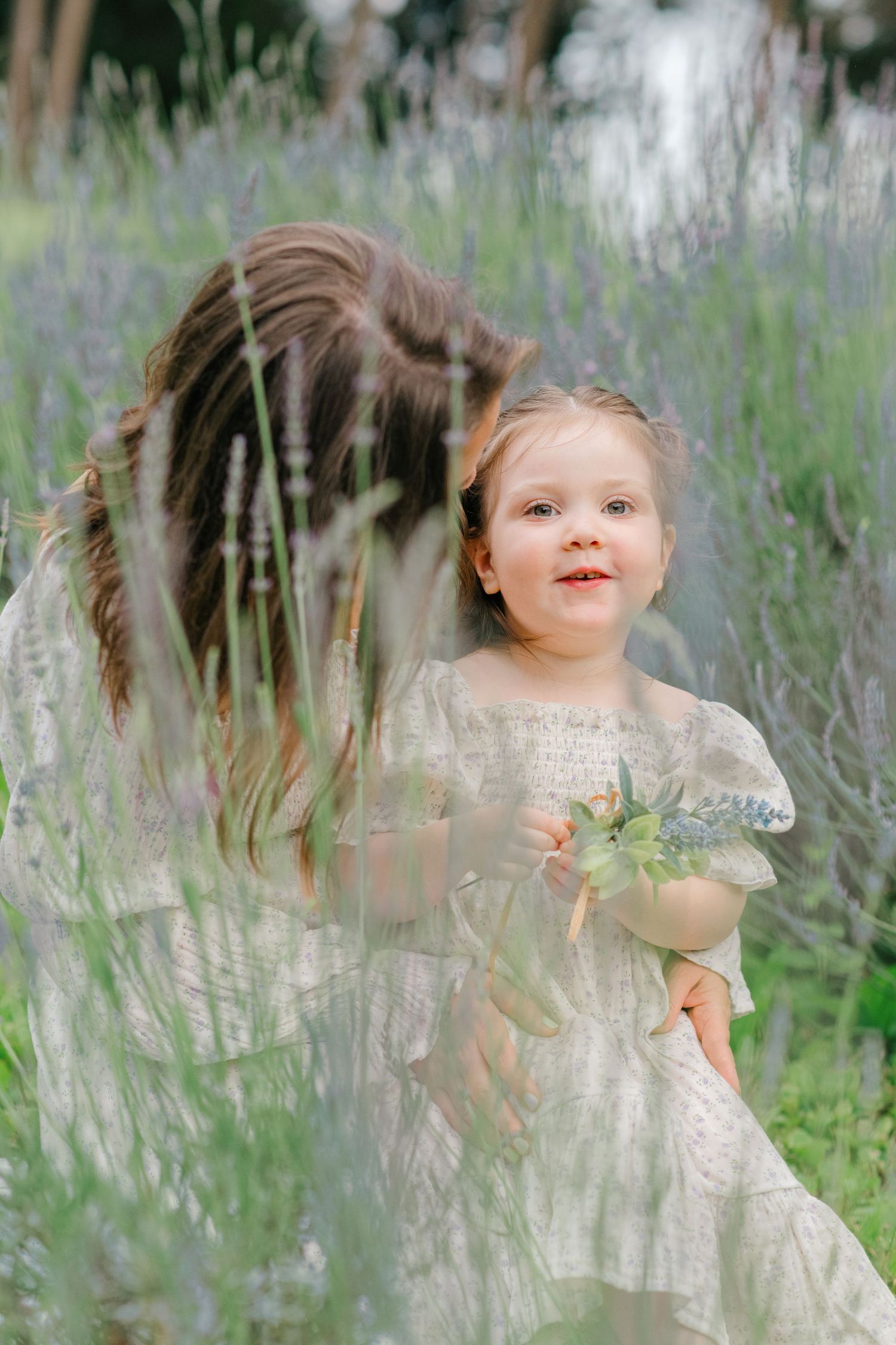 Young girl in vintage dress photographed through lavender flowers in a garden.