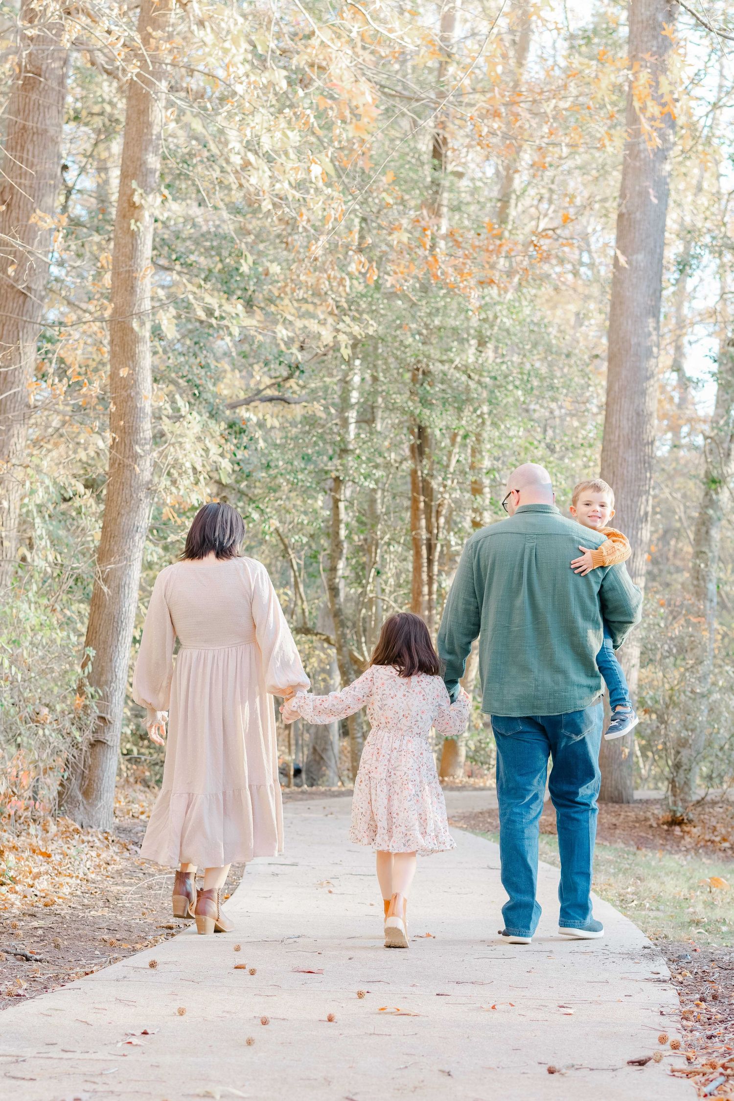 Family mini session in fall of four people walking on a tree-lined path in autumn, parents holding children's hands from behind.