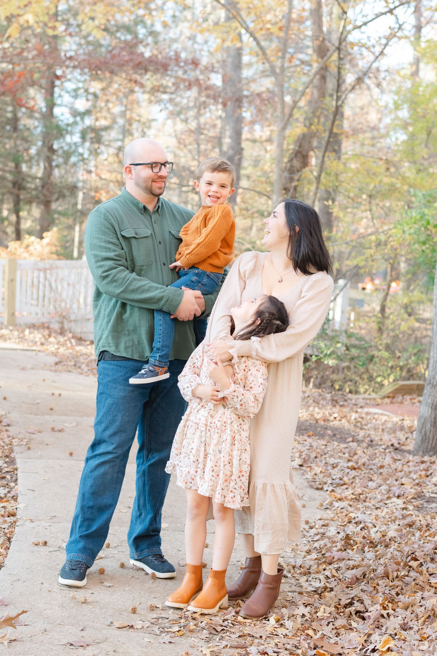 Family of four poses outdoors in autumn, parents holding young children among fall foliage in warm seasonal attire.