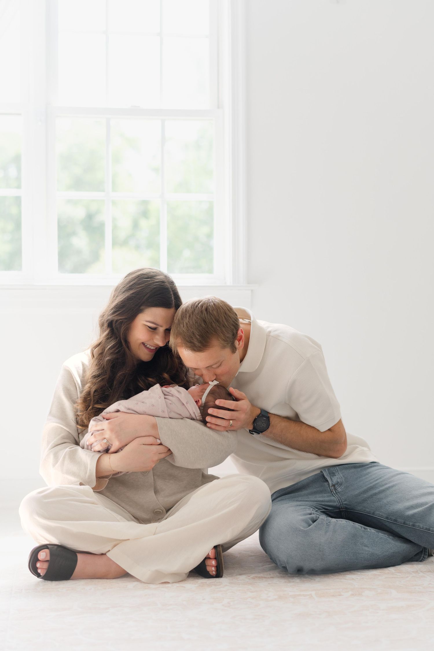 A couple sits on the floor holding and kissing their newborn baby near a bright window.