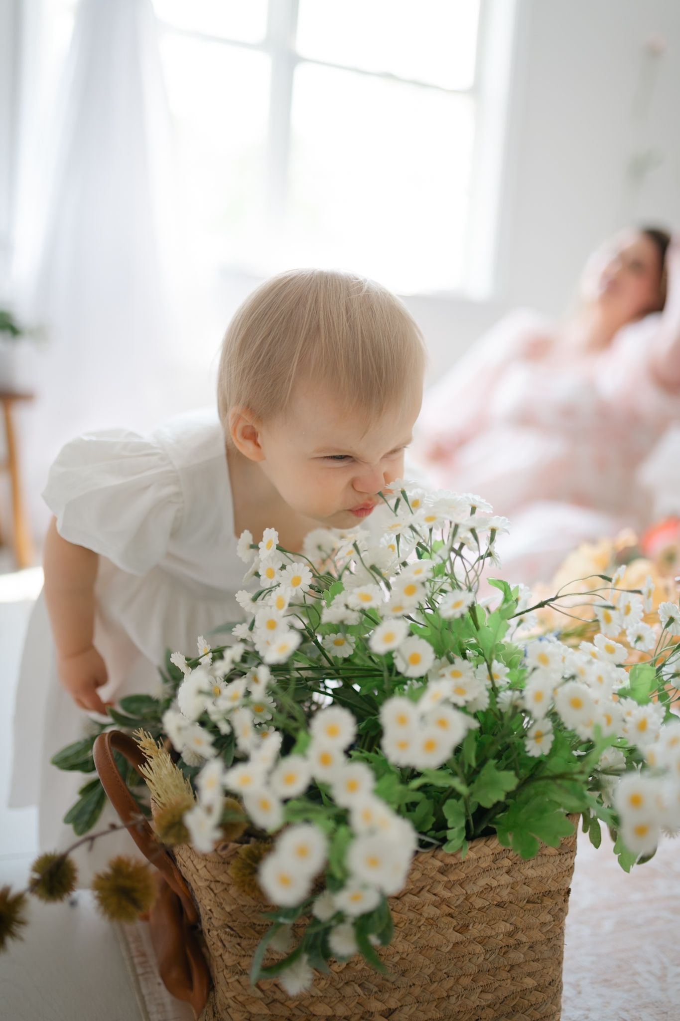 A small child leans over to smell white flowers arranged in a rustic woven basket in Richmond newborn photography studio
