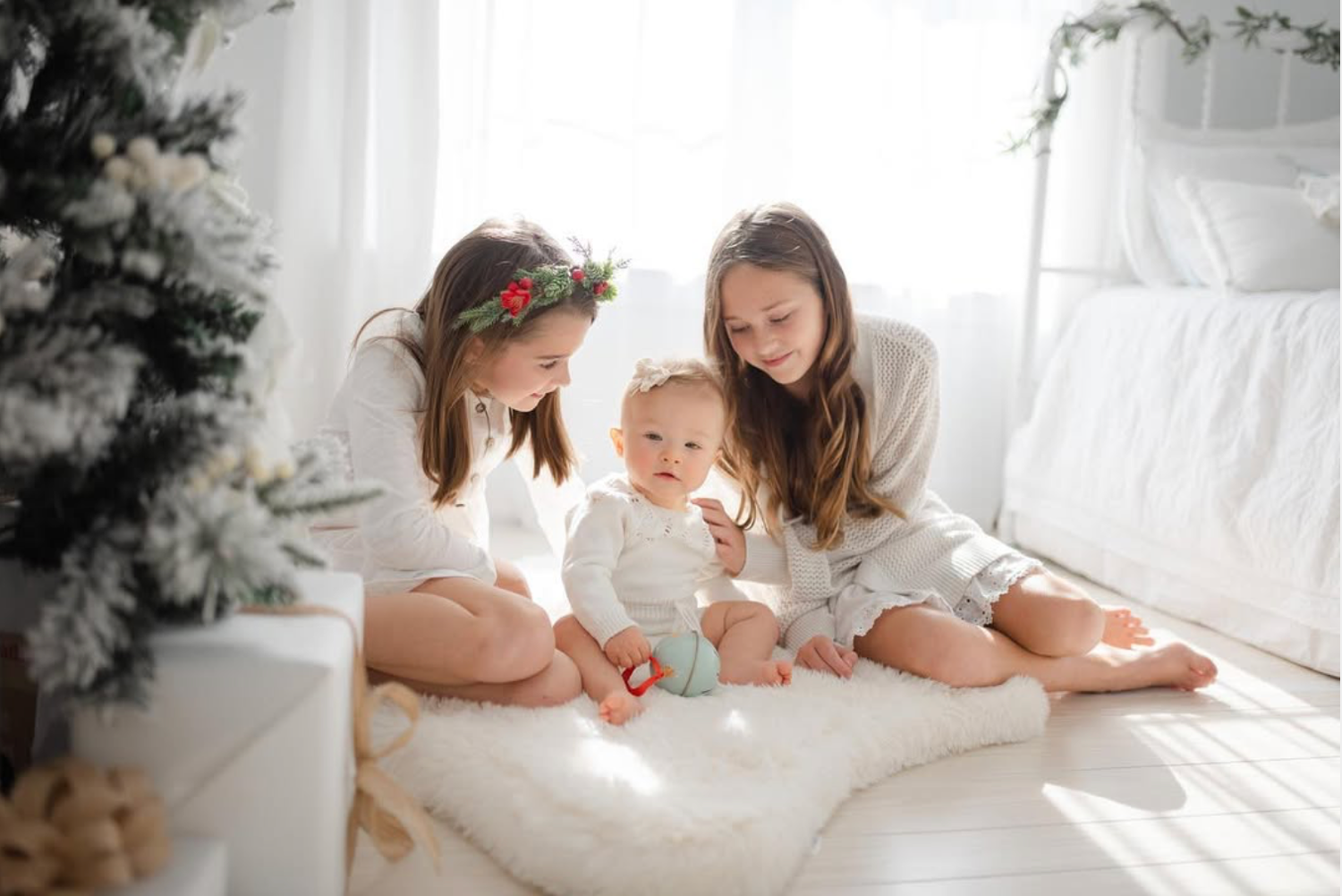 Two sisters in white outfits sit with a baby on a fur rug near a Christmas tree in a bright, decorated room.
