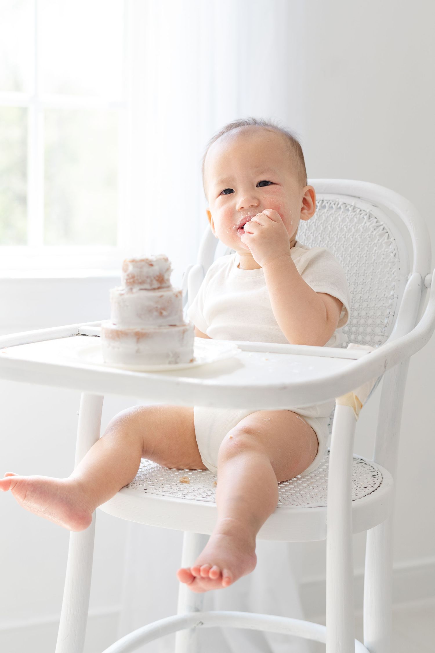 Happy baby sitting in white high chair eating first birthday smash cake, smiling with frosting on hands.