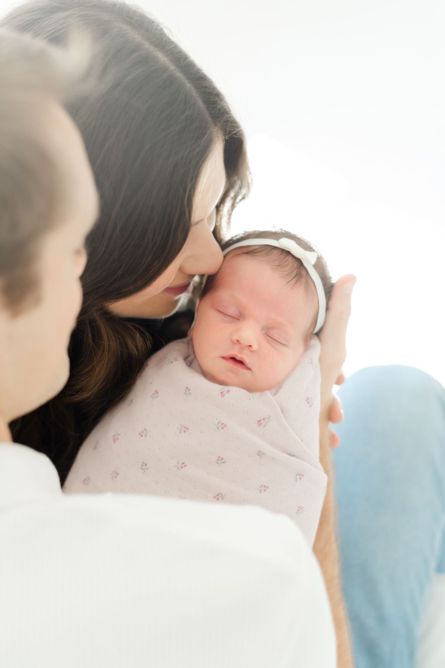 A mother tenderly kisses her sleeping swaddled newborn baby girl wearing a white headband.
