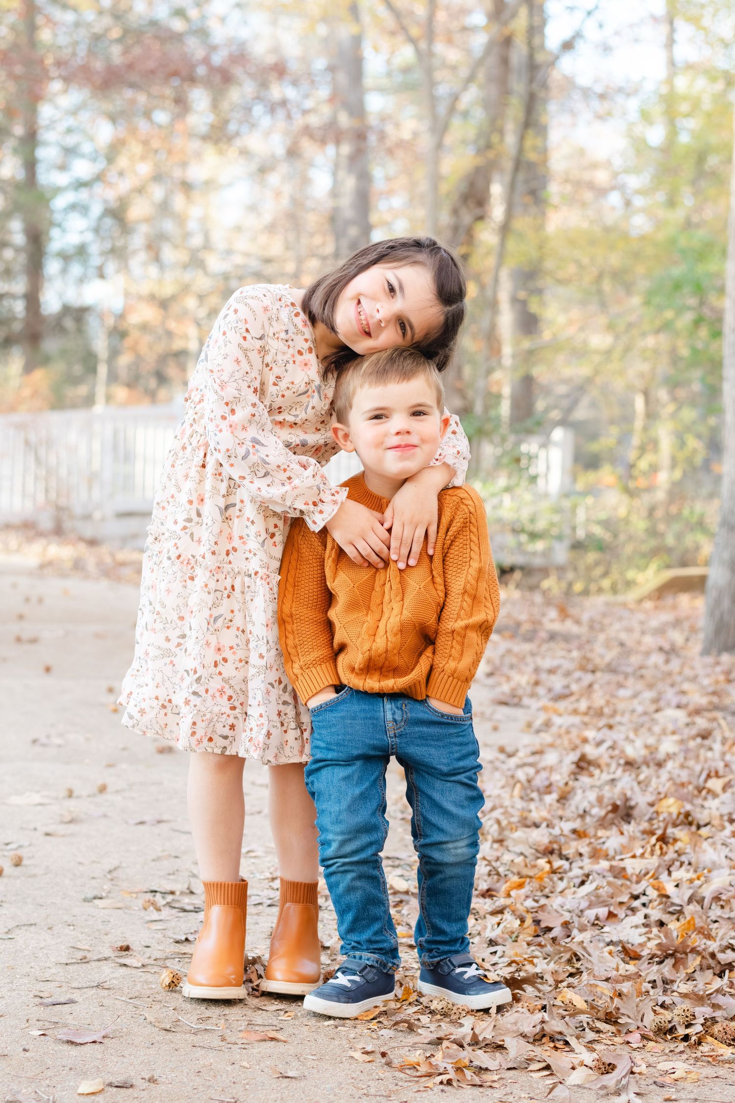 A girl in a floral dress hugs a boy in an orange sweater and jeans outdoors among autumn fallen leaves.