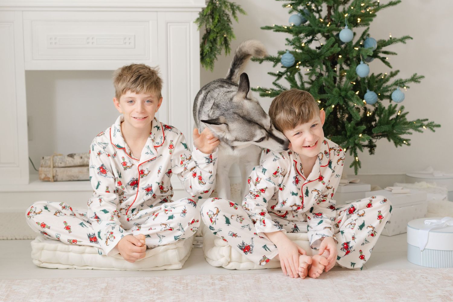 Two boys in matching Christmas pajamas sit on a white rug beside a decorated tree, playing with a gray rabbit.