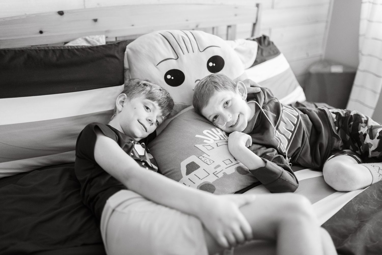 Two young boys smiling and cuddling with a large stuffed animal pillow on a bed in a black and white photo.