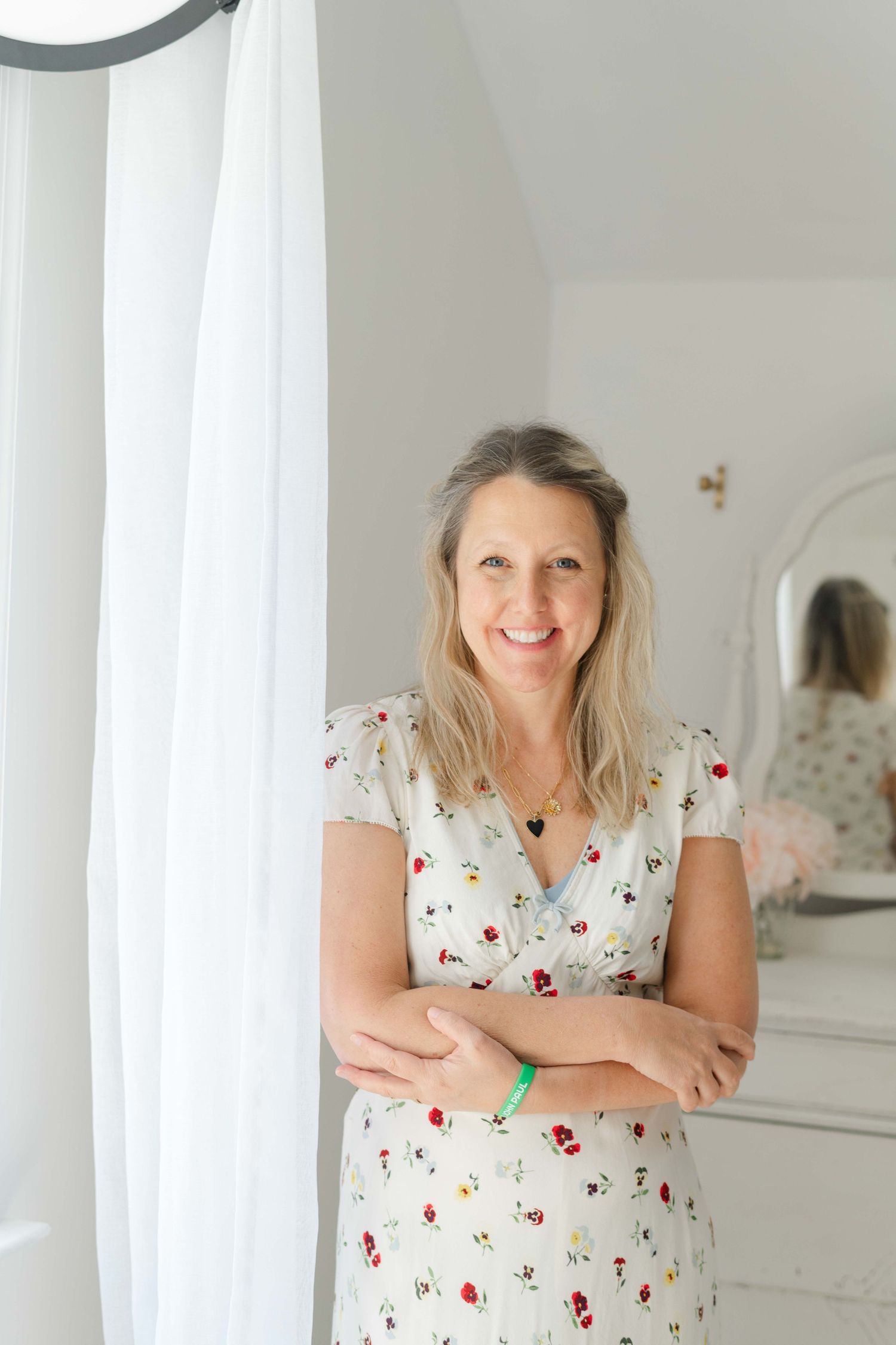 Smiling blonde woman in floral dress stands by white curtain indoors.
