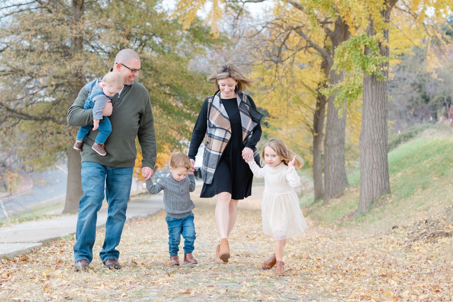 Family of four walking outdoors in autumn park with colorful fall foliage, parents holding children's hands, smiling together.