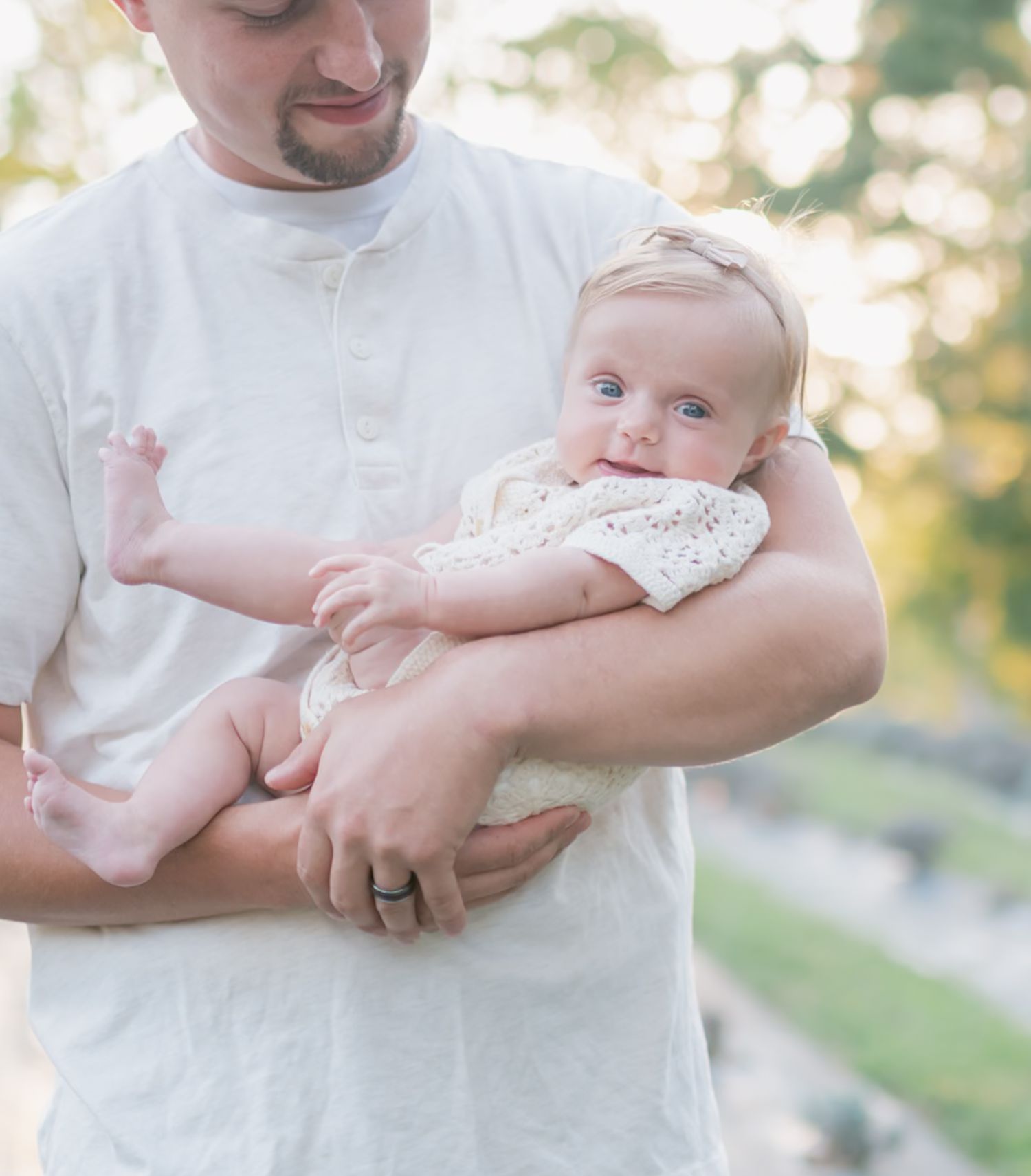 Parent gently cradles baby in white outfit during outdoor golden hour photo session.
