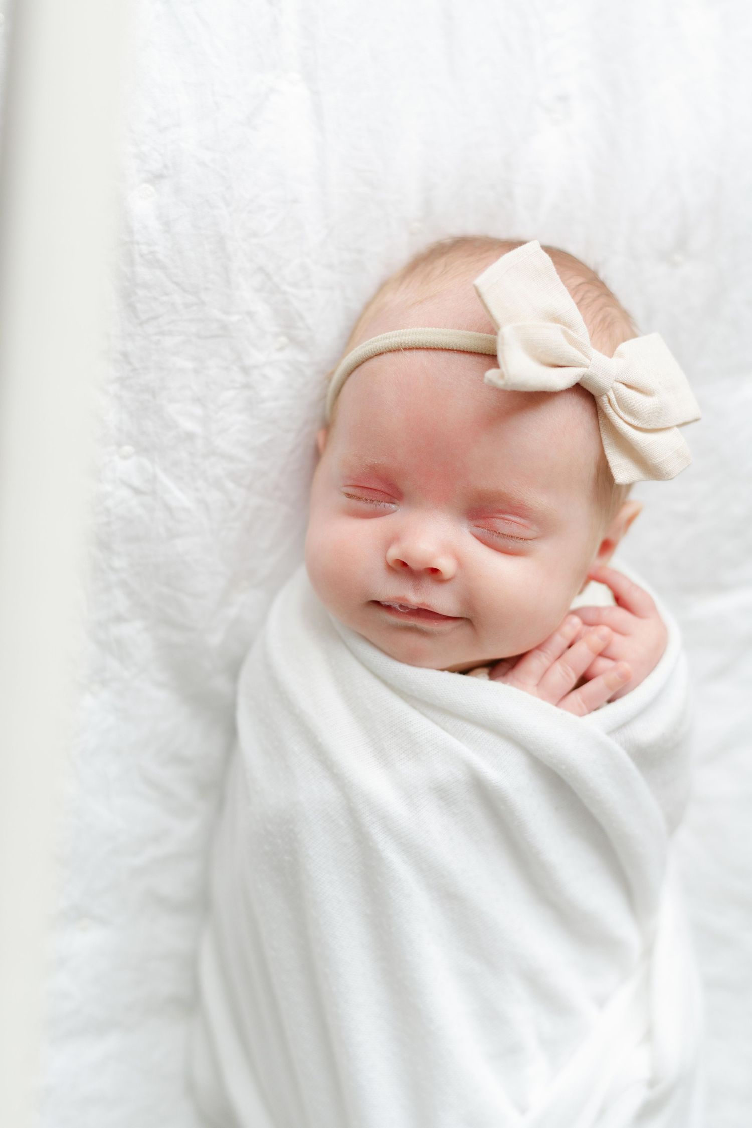 A peaceful sleeping newborn baby swaddled in white with a delicate cream bow headband resting on soft bedding.