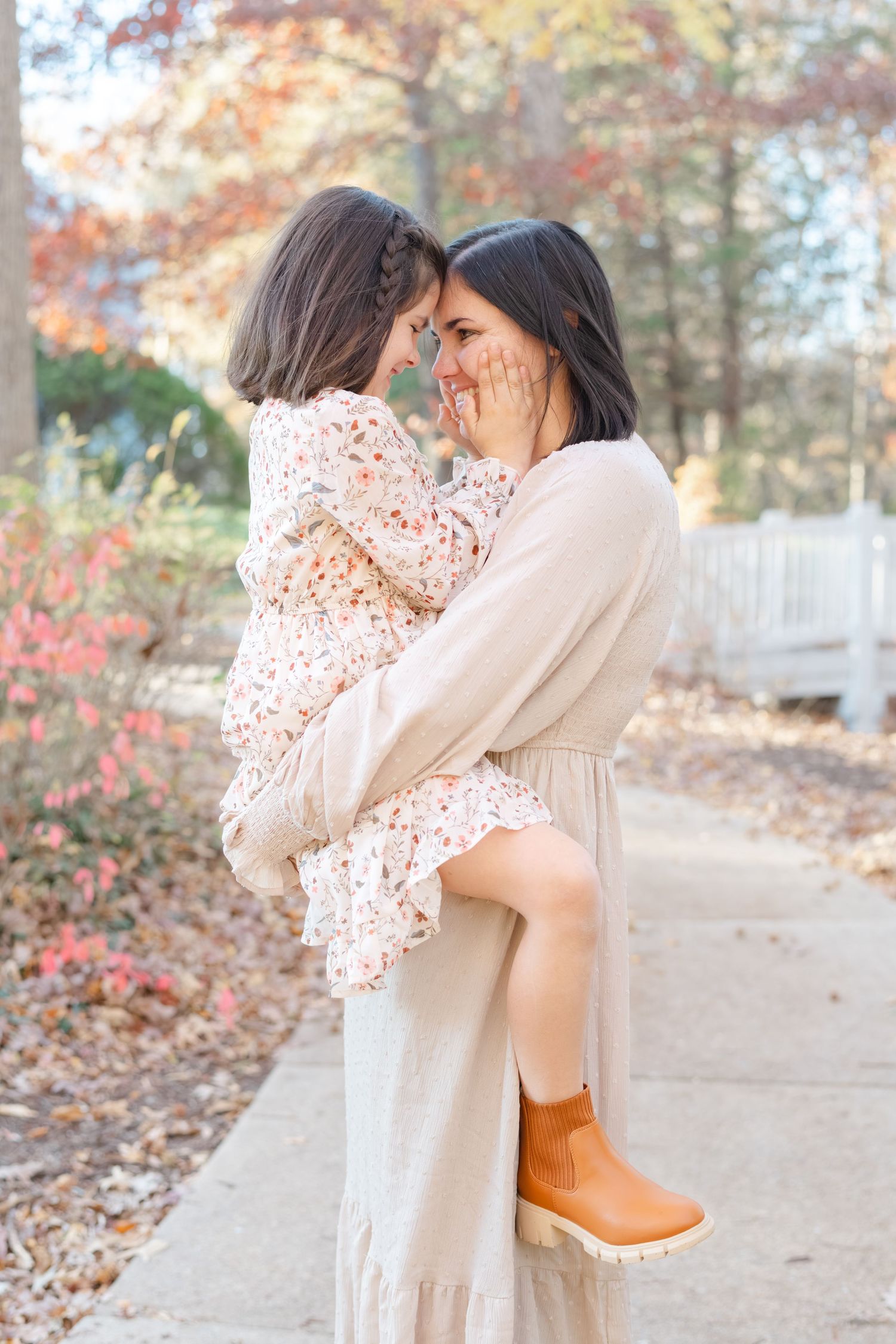 A mother holds her young daughter outdoors in fall, both wearing floral and cream outfits, touching foreheads affectionately.