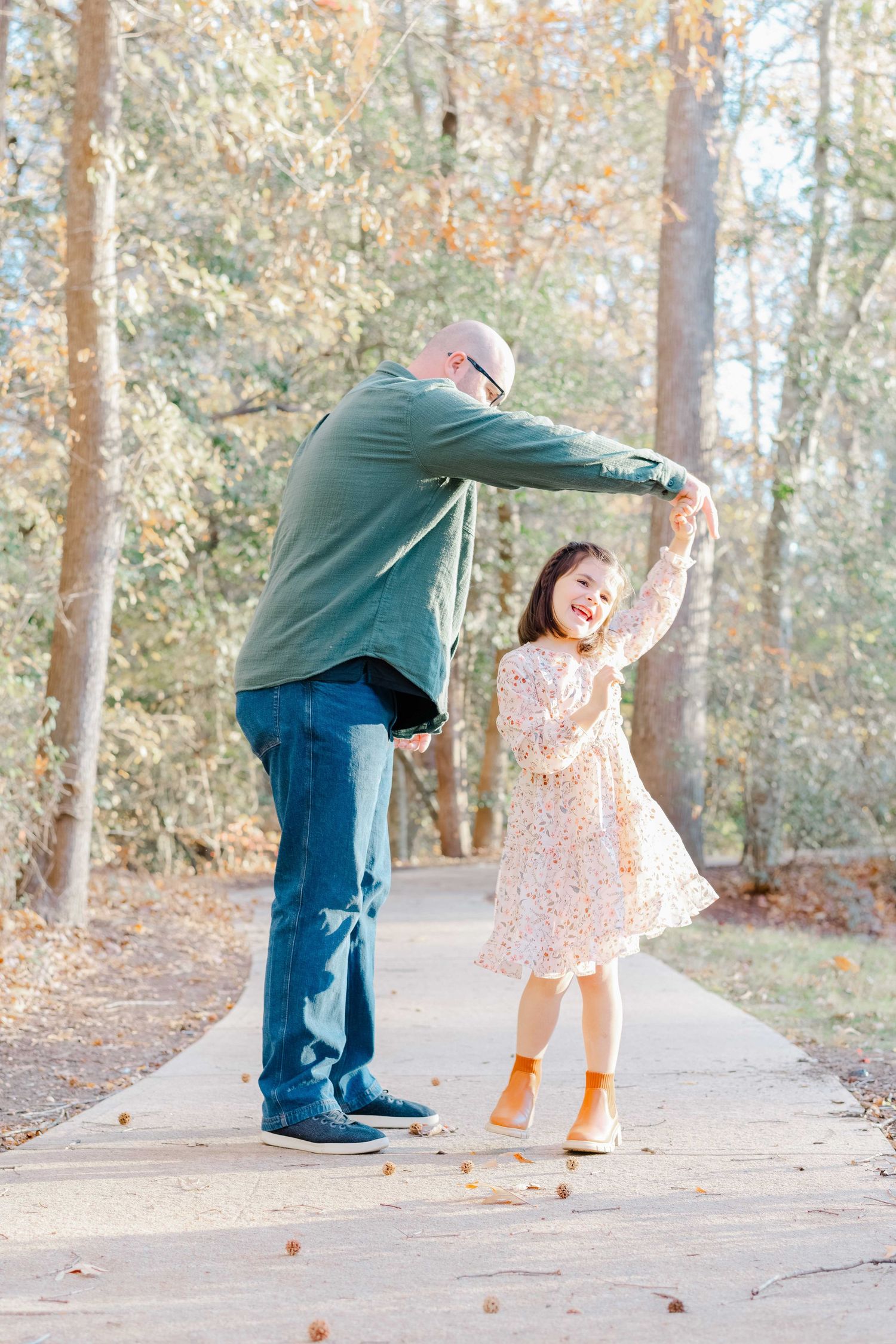 A father twirls his young daughter on a sunlit autumn path surrounded by fall foliage trees.