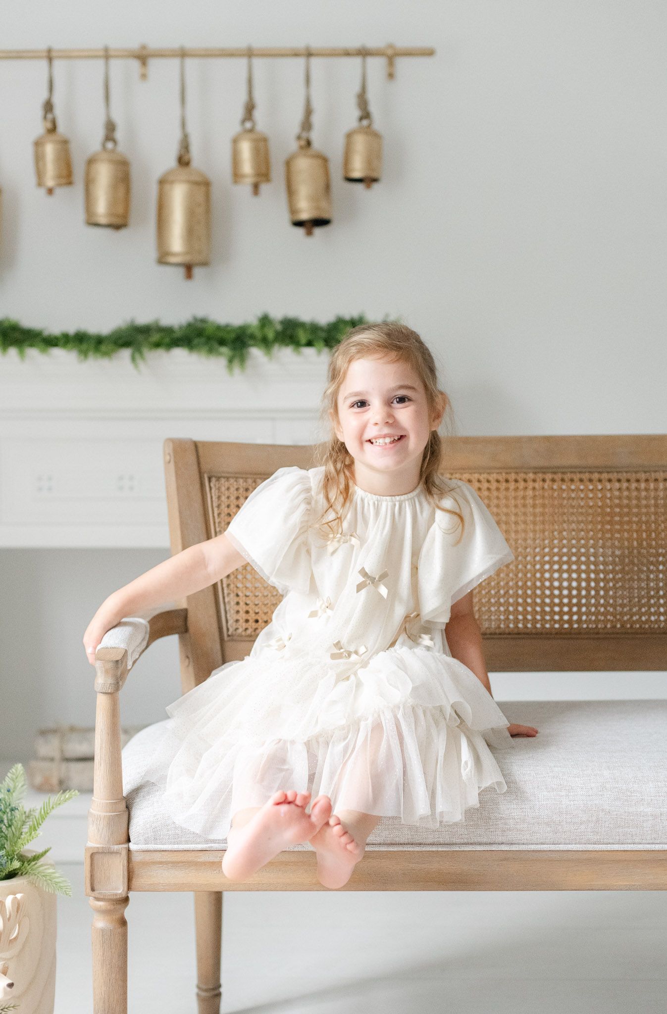 A white dress sits on a wooden bench with brass bells hanging on the wall and greenery garland decorations.