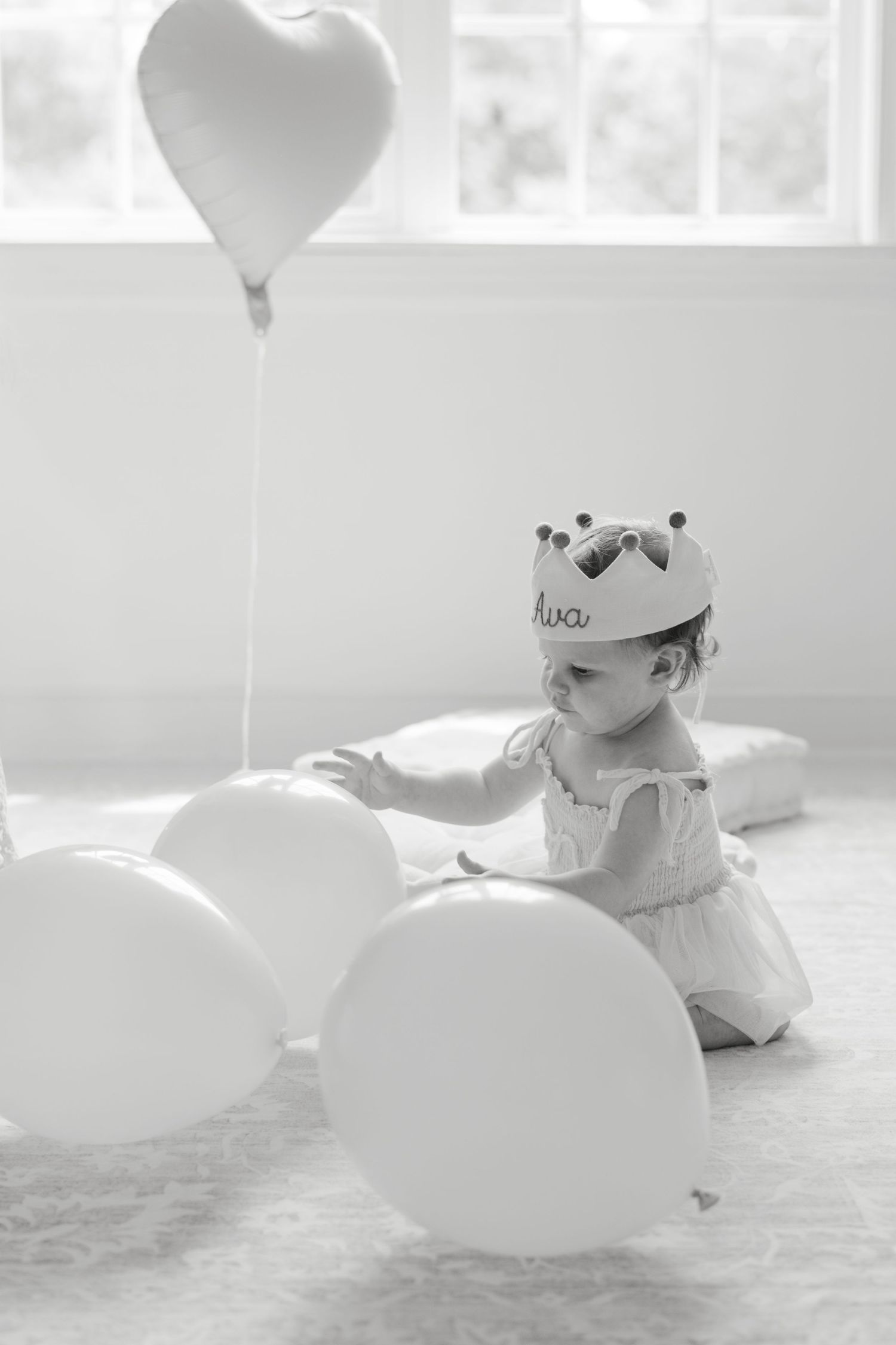 Black and white photo of baby girl wearing a crown, playing with white balloons including a heart-shaped one.