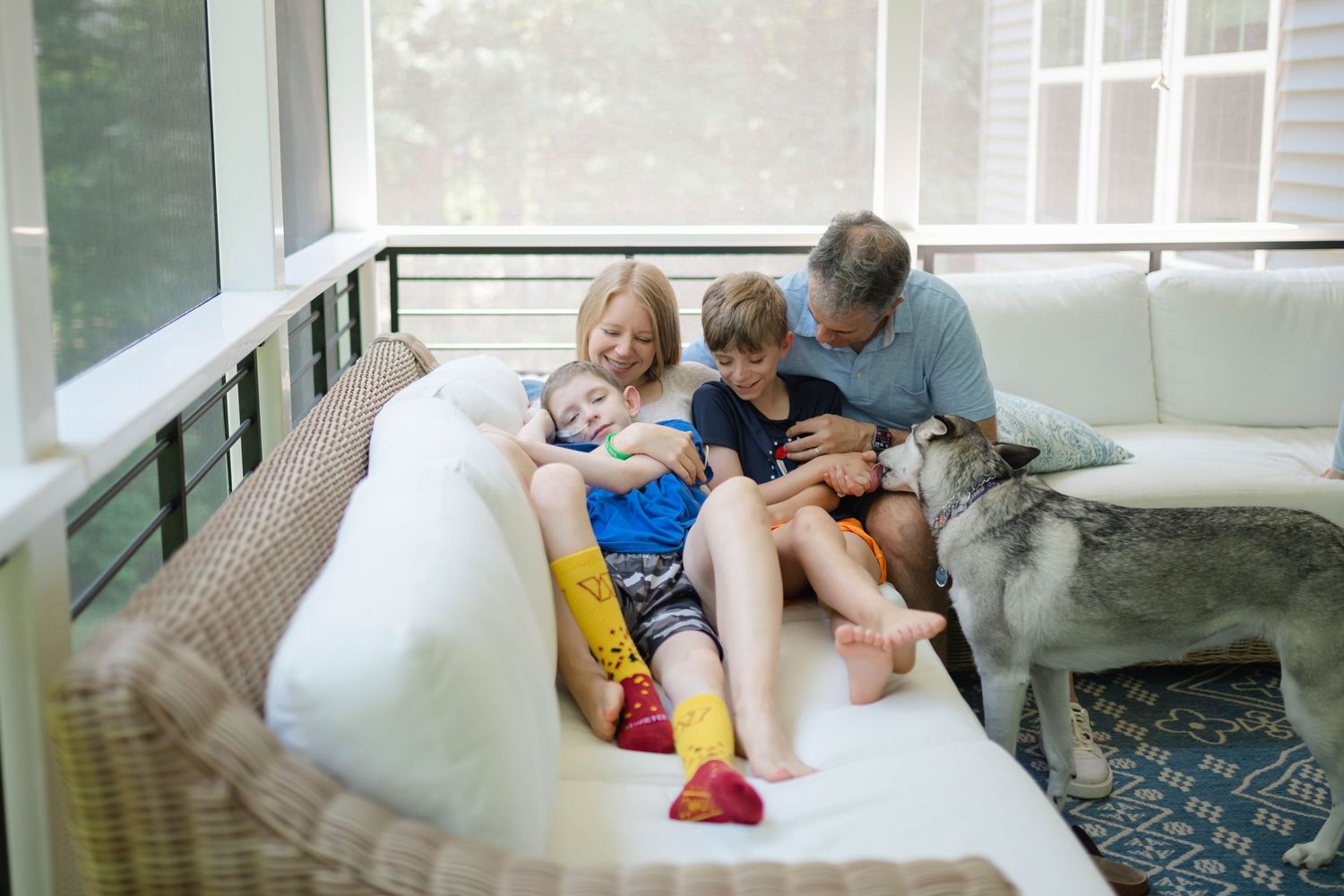 Family with two kids and a husky dog relaxing together on a wicker porch sofa in a screened sunroom.