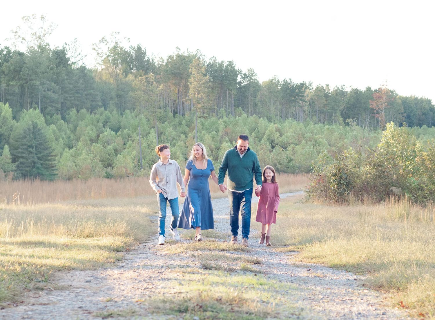 A group of people walking on a gravel path through a scenic autumn meadow with pine trees in the background.