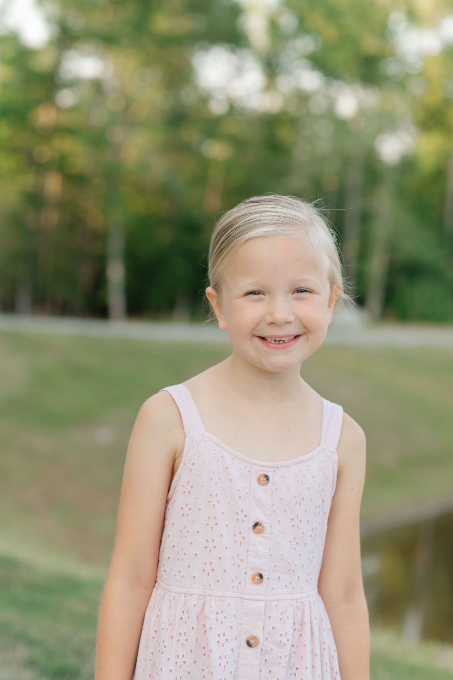 Young blonde girl in pink sundress smiling outdoors with green trees and pond in background.