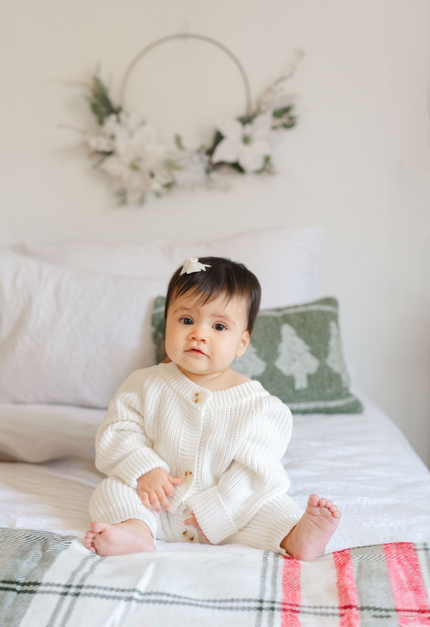 Adorable baby in cream knit romper sitting on bed with plaid blanket and floral hoop wreath backdrop.