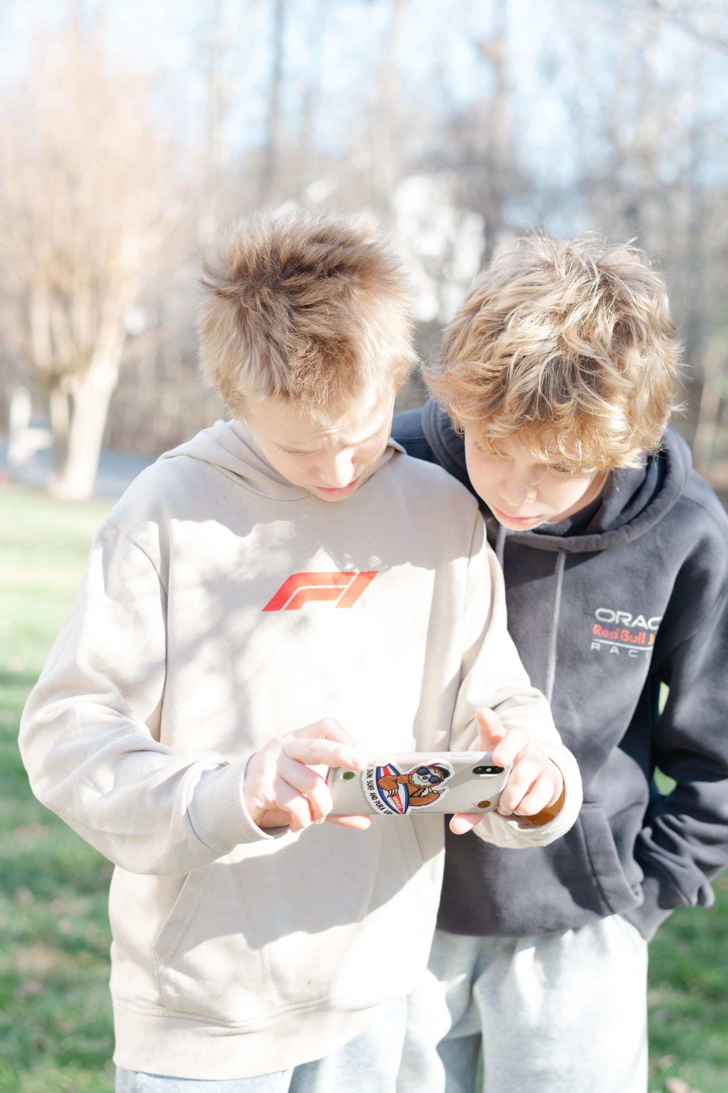 Two people in sweatshirts examine something together outdoors on a sunny day with trees in the background.