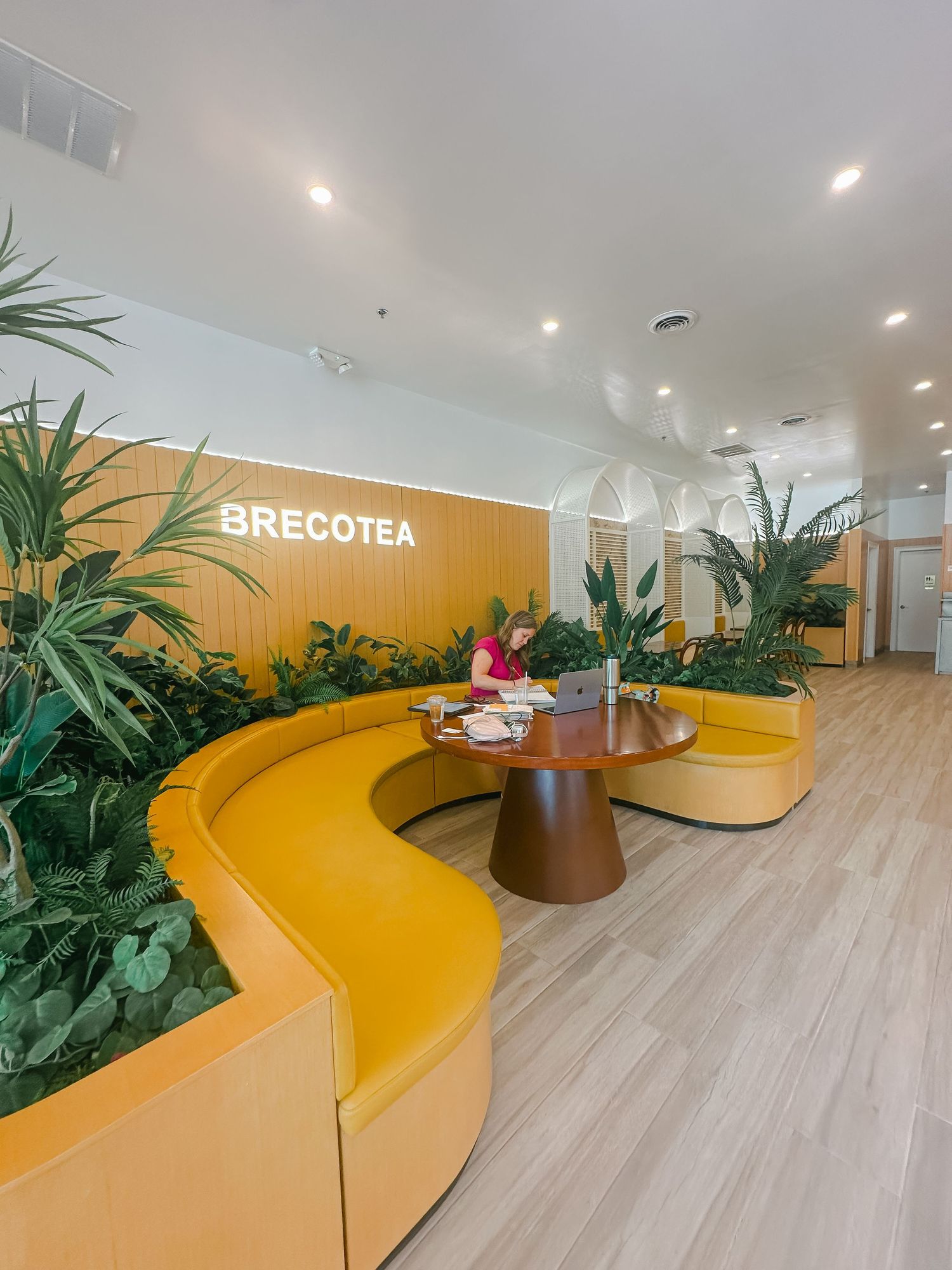 Modern office lounge area with curved yellow seating and tropical plants against white walls.