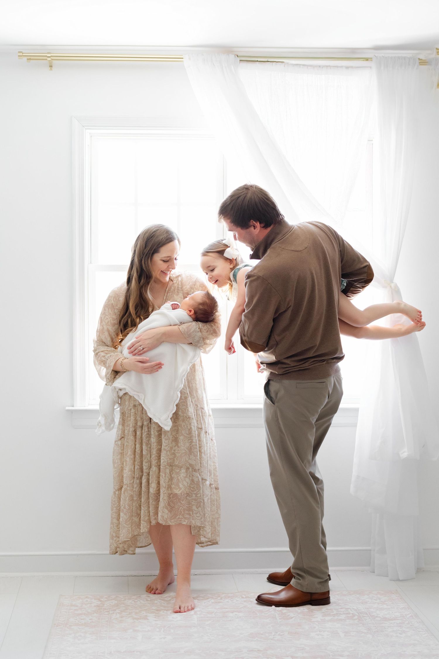 A family of four poses by a bright window, parents lovingly holding their newborn and toddler in a soft, airy white room.