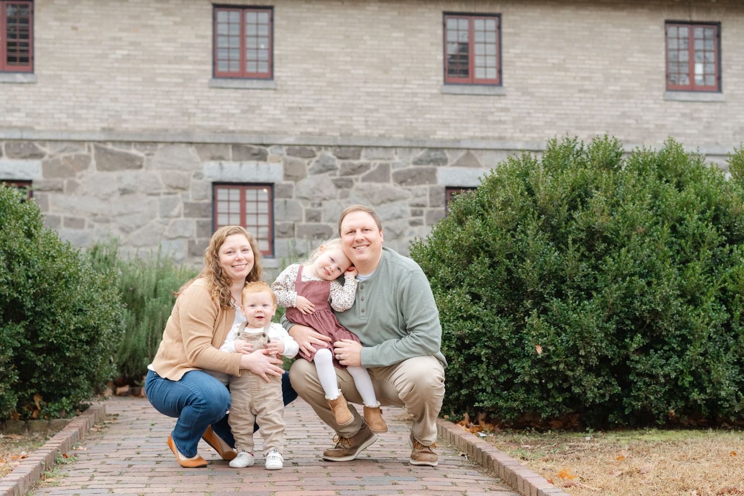 Family of four posing outdoors in front of a stone building, parents crouching with two young children between green shrubs.