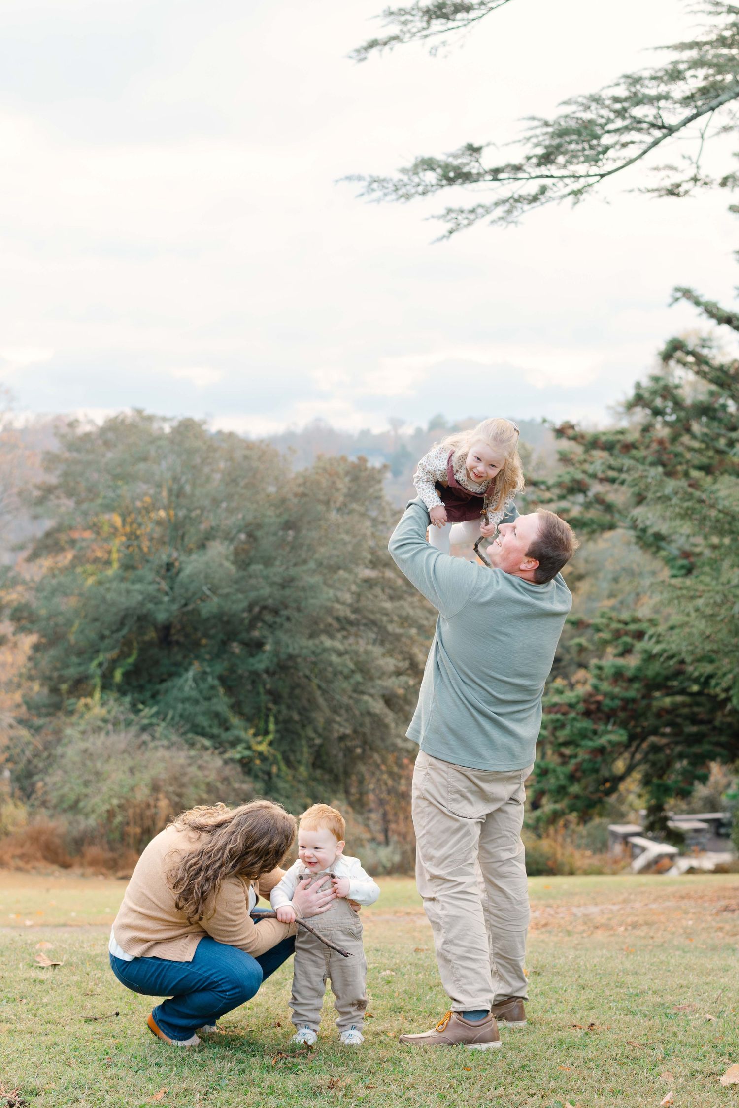 Family of four enjoying outdoor fall photos, with dad lifting child and mom crouching with toddler in scenic park.