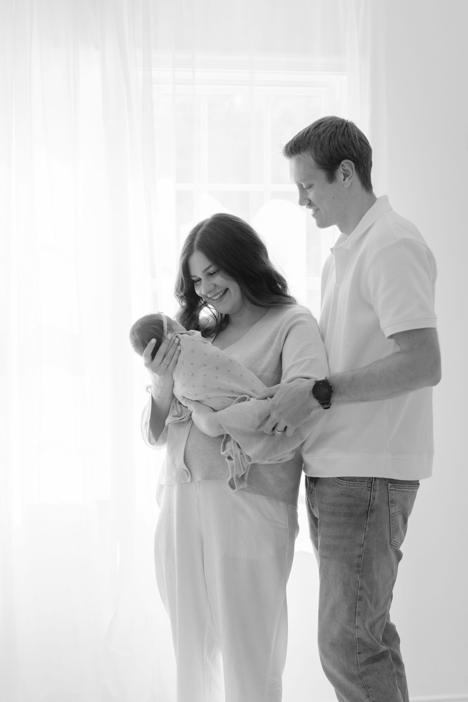 Black and white photo of parents smiling at their newborn baby by a bright window.