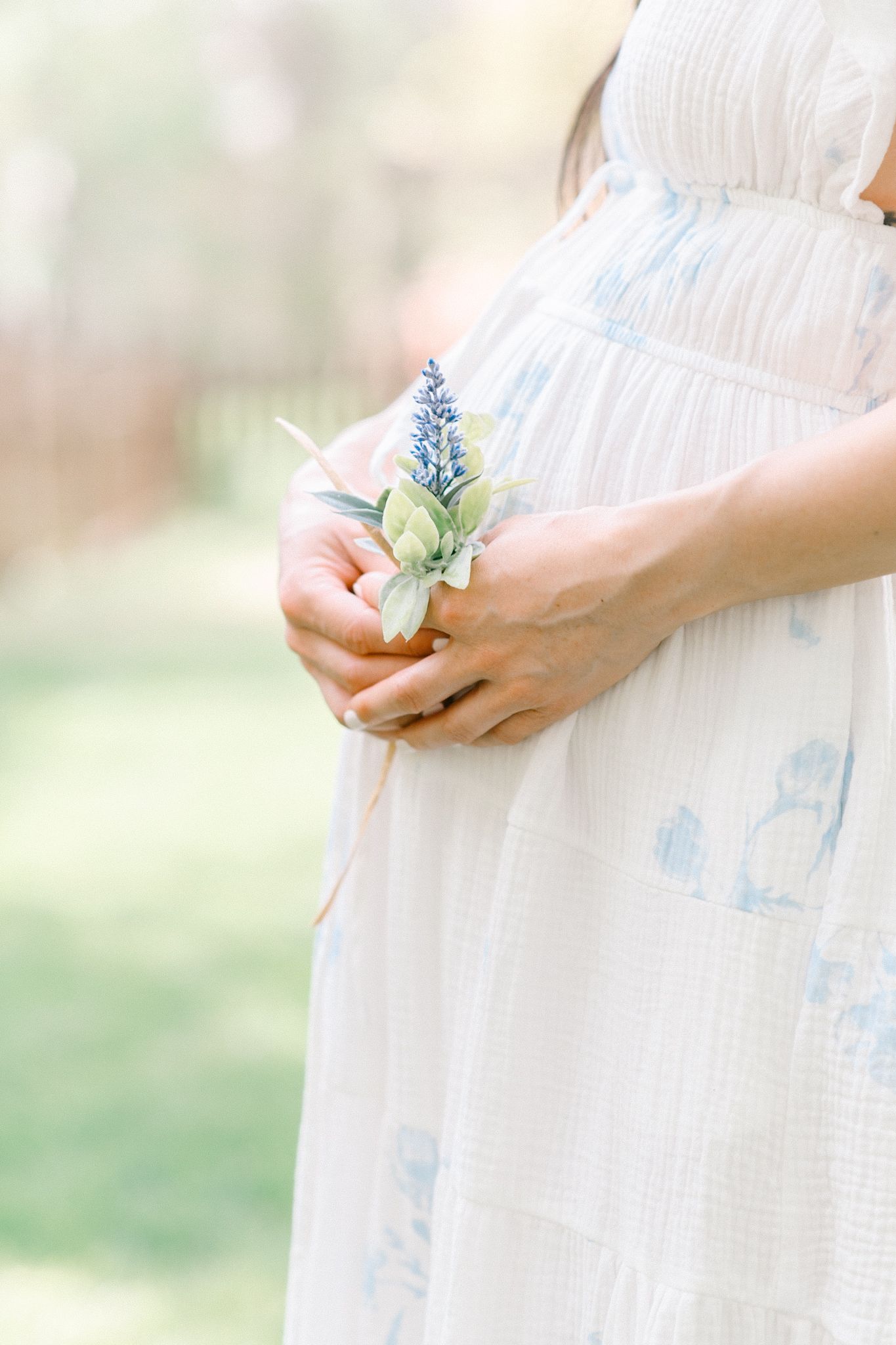 Delicate hands holding a small blue flower against a white dress with soft blue floral pattern in dreamy light.