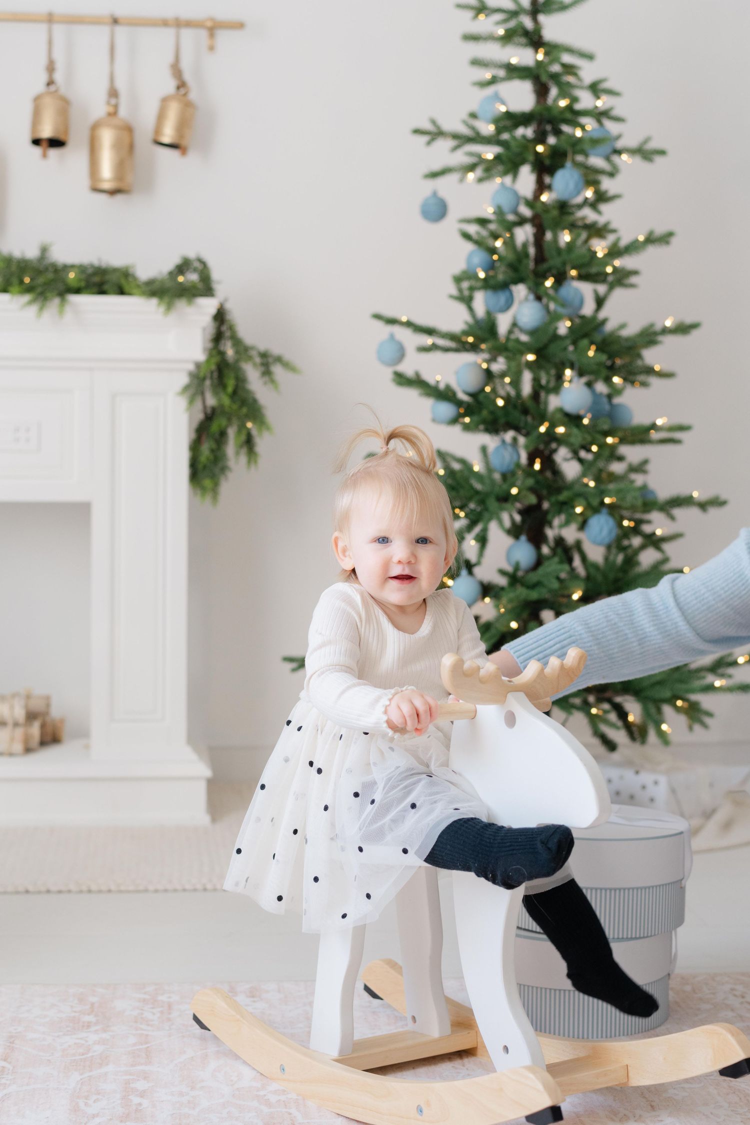 Toddler girl in white dress riding a wooden rocking reindeer toy near a decorated Christmas tree.