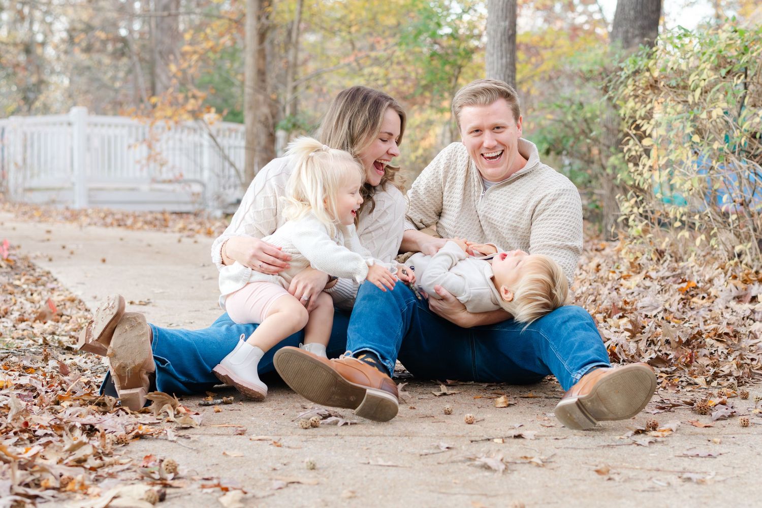 Happy family of four sitting on a leaf-covered path in autumn, laughing and playing together outdoors.