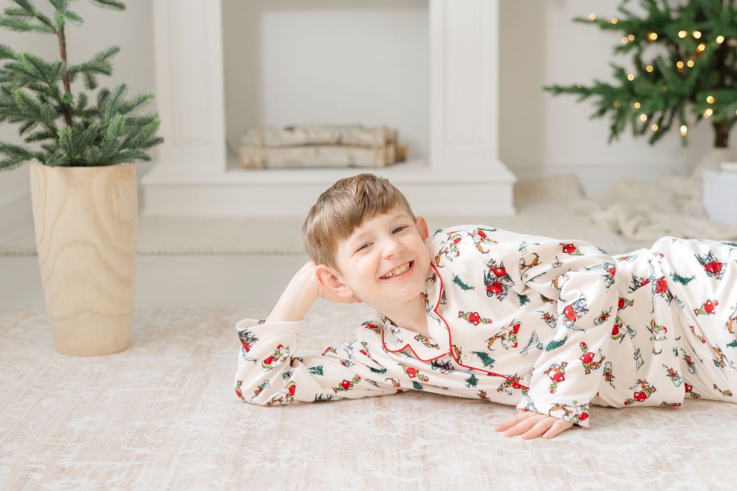 Smiling boy in holiday pajamas lying on floor near Christmas trees in festive white room.
