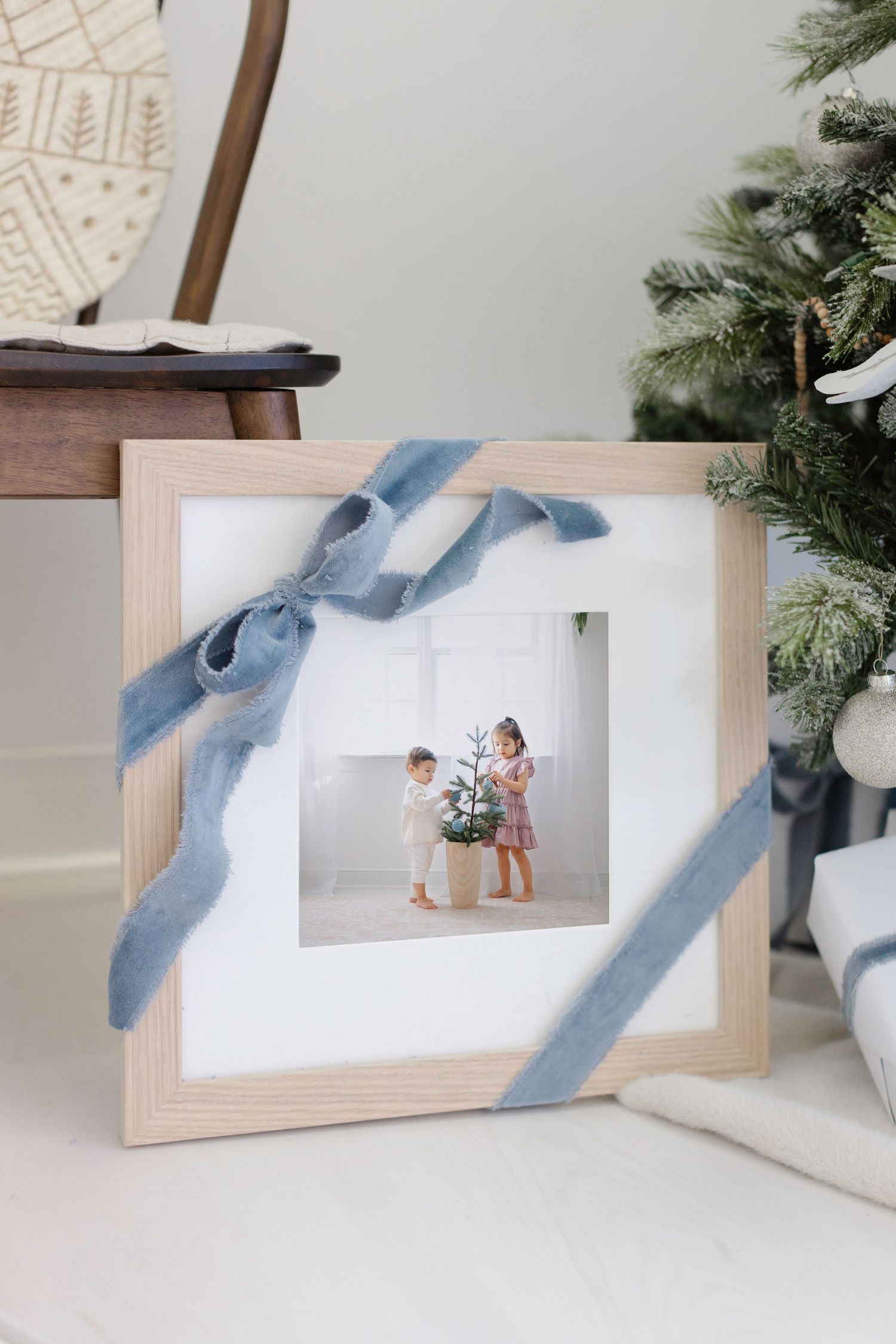 Wooden photo frame with blue velvet ribbon bow displaying children with Christmas tree, placed near holiday decorations.