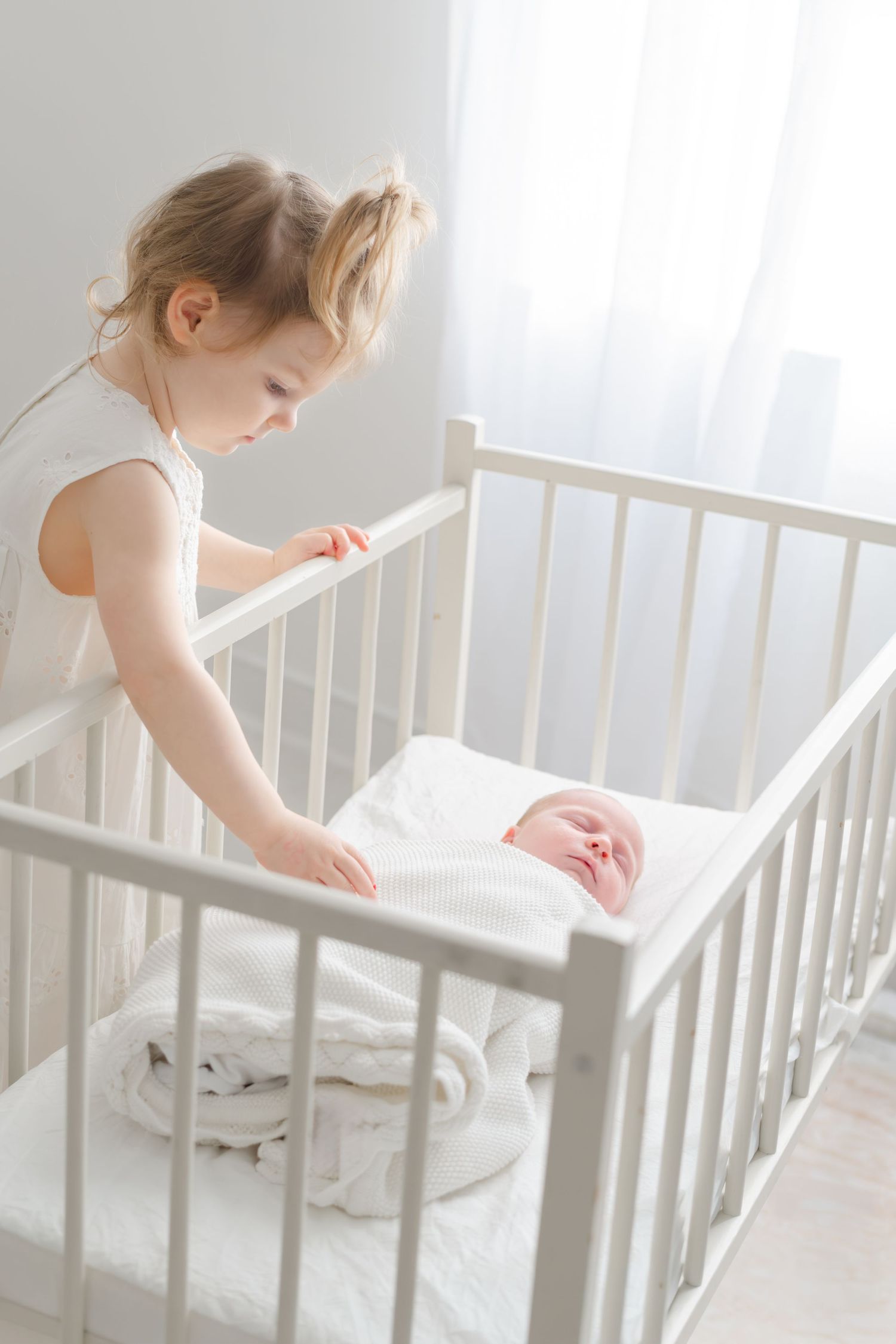 Young girl in white dress leaning over white crib looking at sleeping newborn baby sibling indoors.