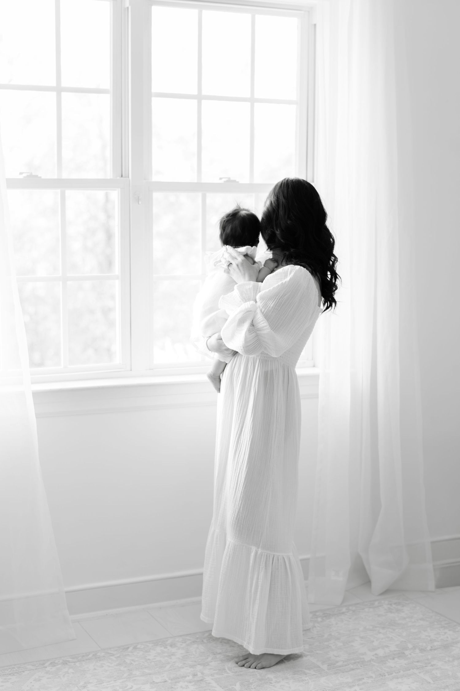 Black and white photo of a mother in a white dress holding her newborn baby by a bright window.