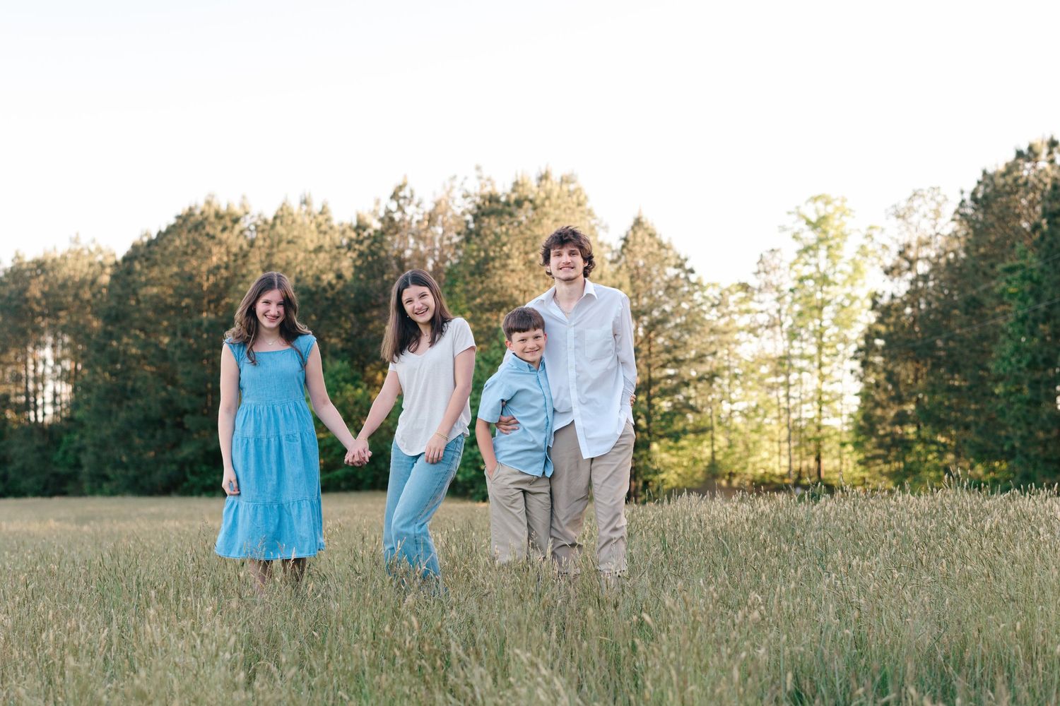 Family of four holding hands walking through a sunny green field with trees in the background.