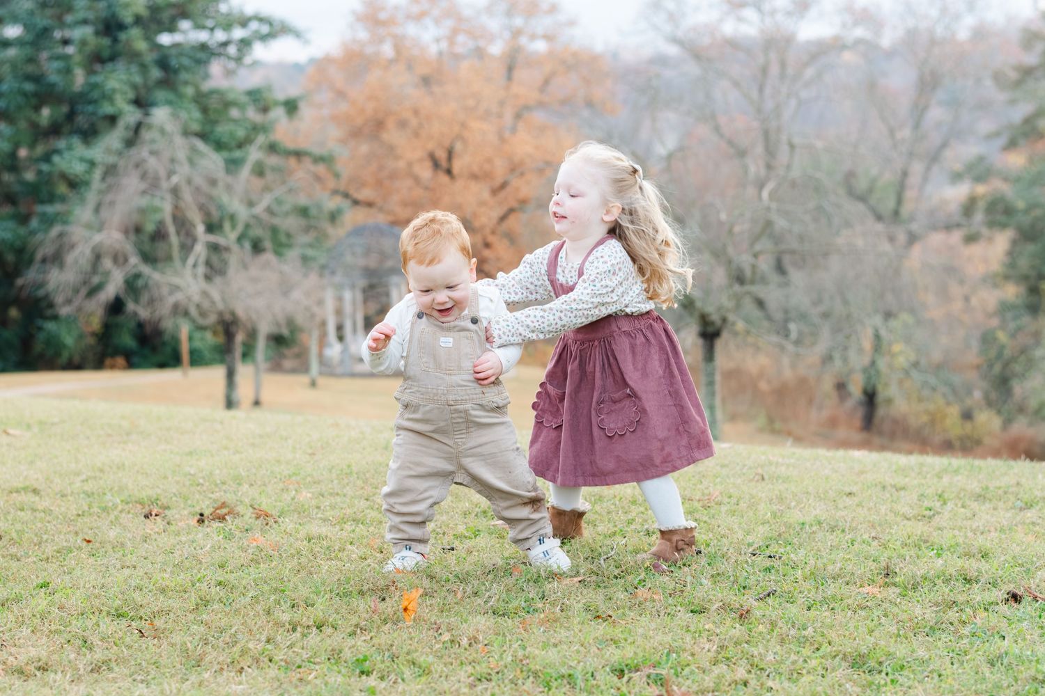 Two young children playing together on a grassy lawn surrounded by autumn trees in a park.