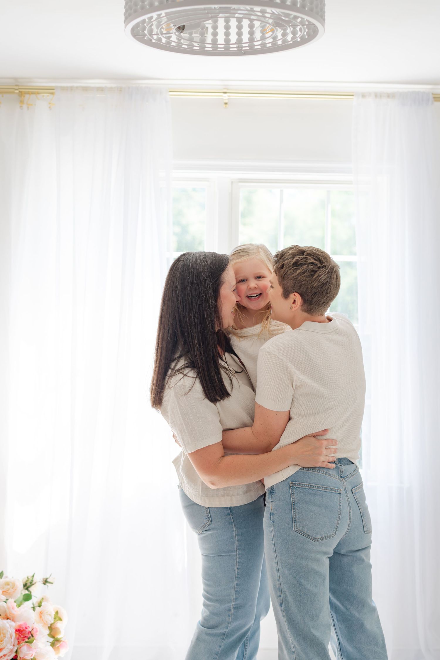 Two women in white tops and jeans share a joyful hug with a laughing blonde child in a bright white room.