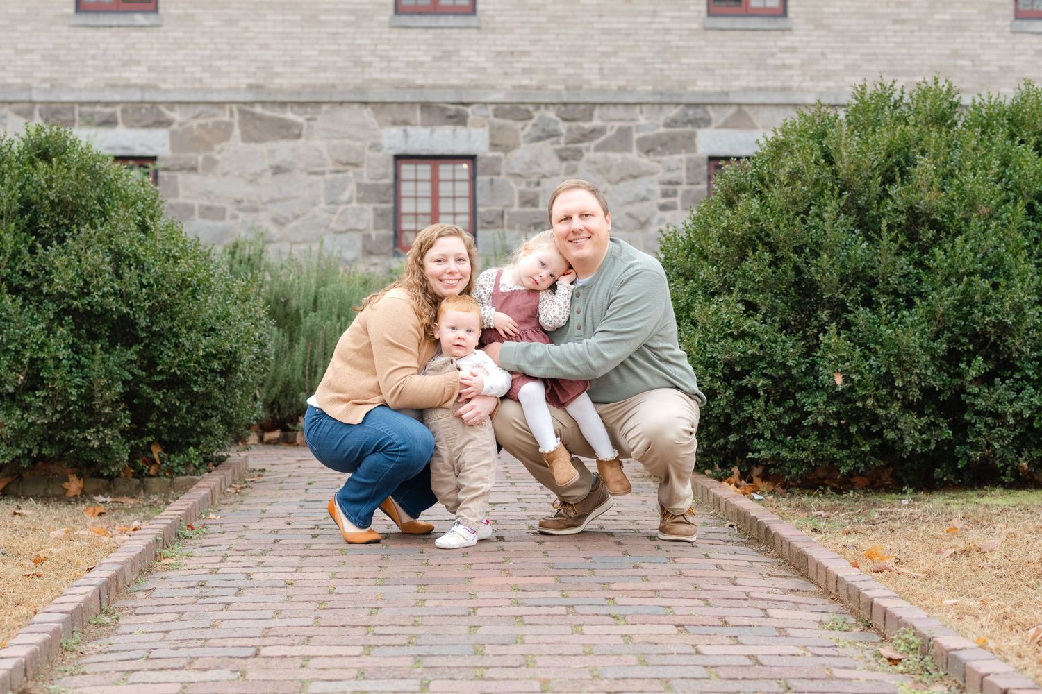 Family of four poses on brick path before stone building, parents crouching with two young children in autumn setting.