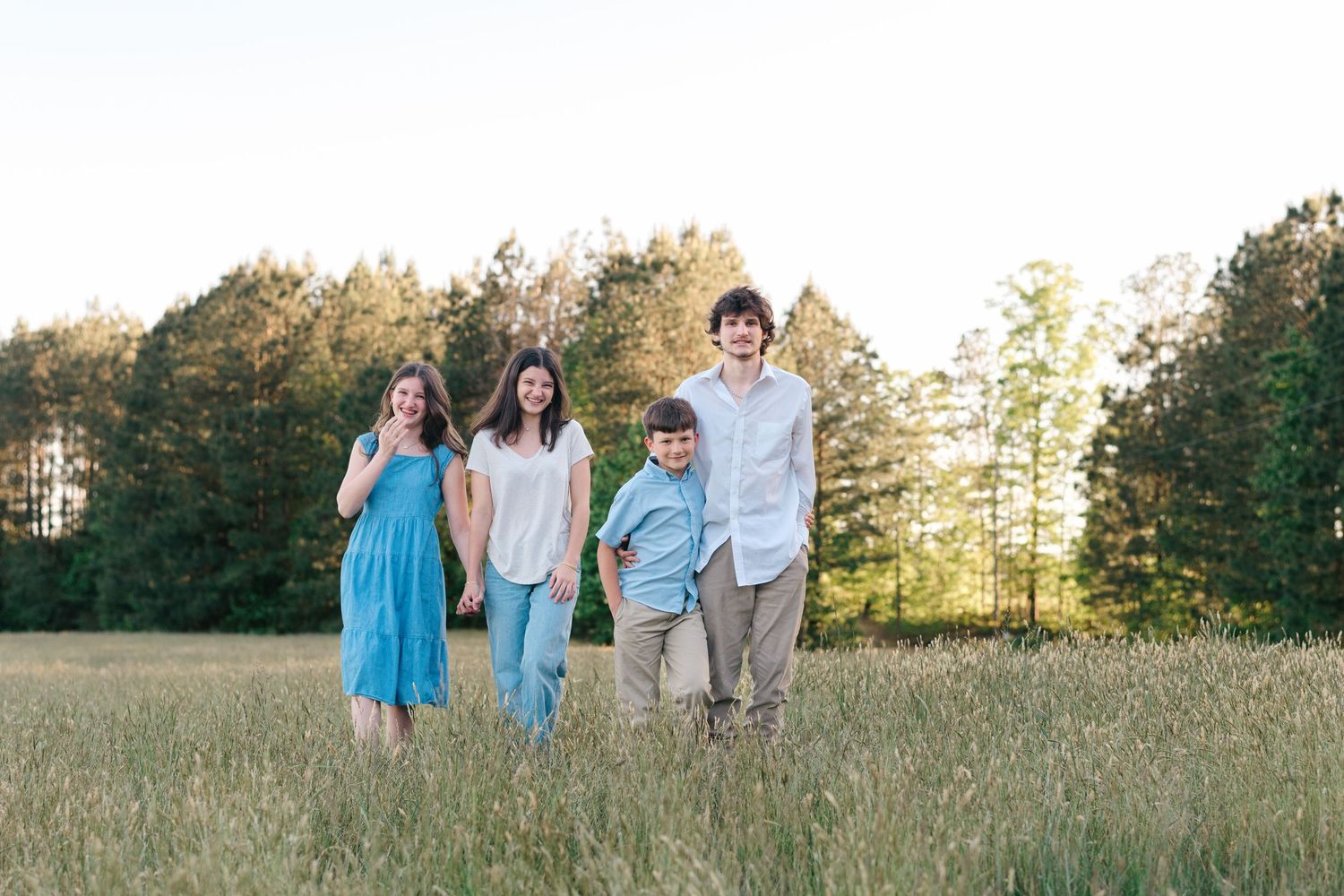 Family of four walking through a sunny meadow with tall grass and trees in the background during golden hour.