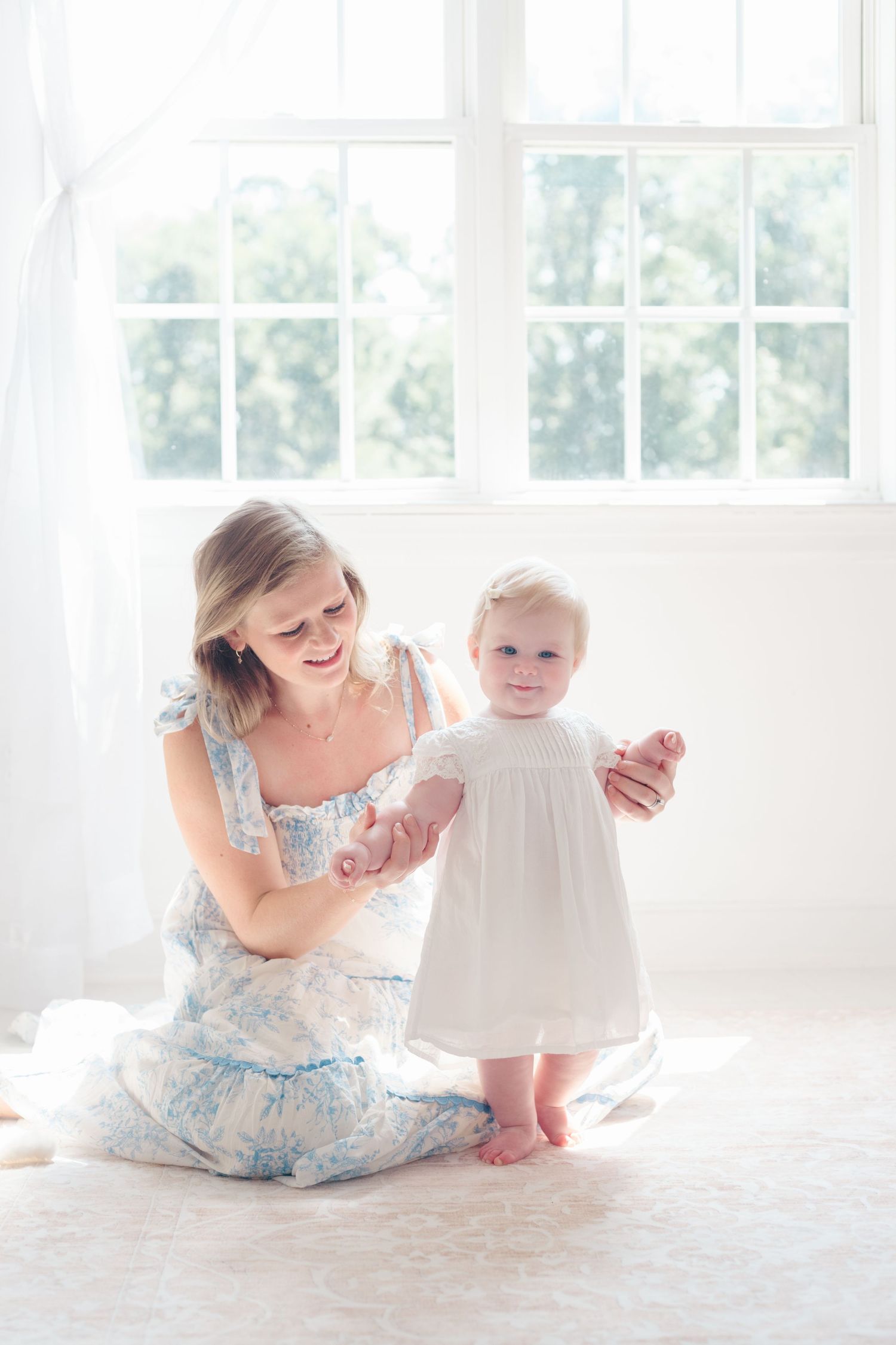 Mother and baby in white and blue floral dresses sit together on light carpet near windows.