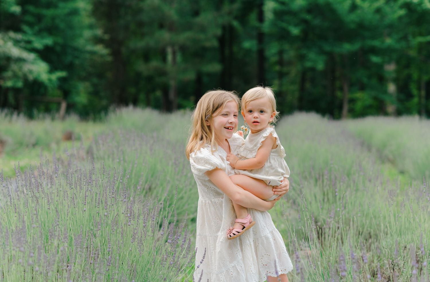 Two young sisters in white dresses pose in a purple lavender field with lush green trees in the background.