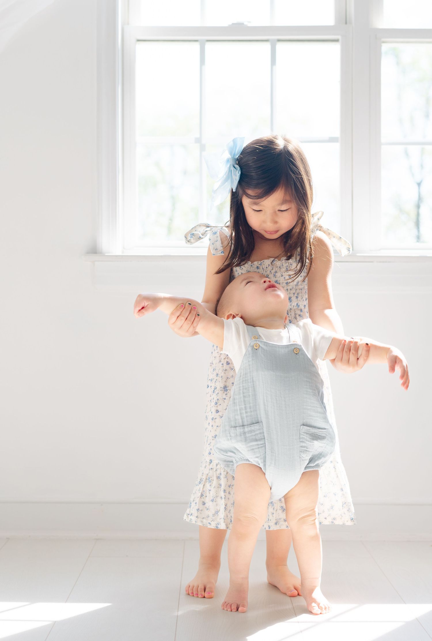 A young girl in a floral dress helps a baby take first steps near a bright window.