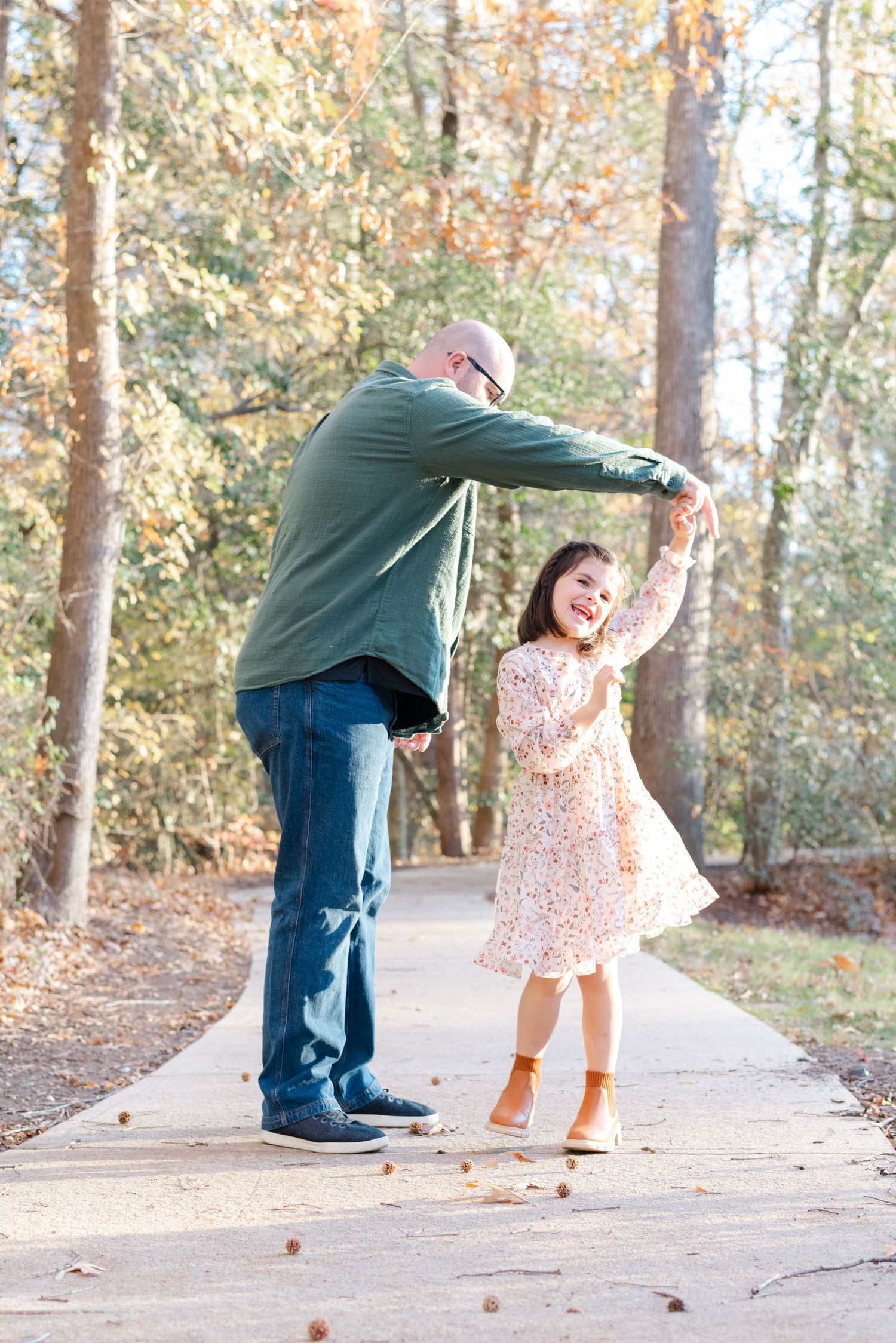 A father twirls his smiling young daughter on a park path surrounded by colorful autumn trees.